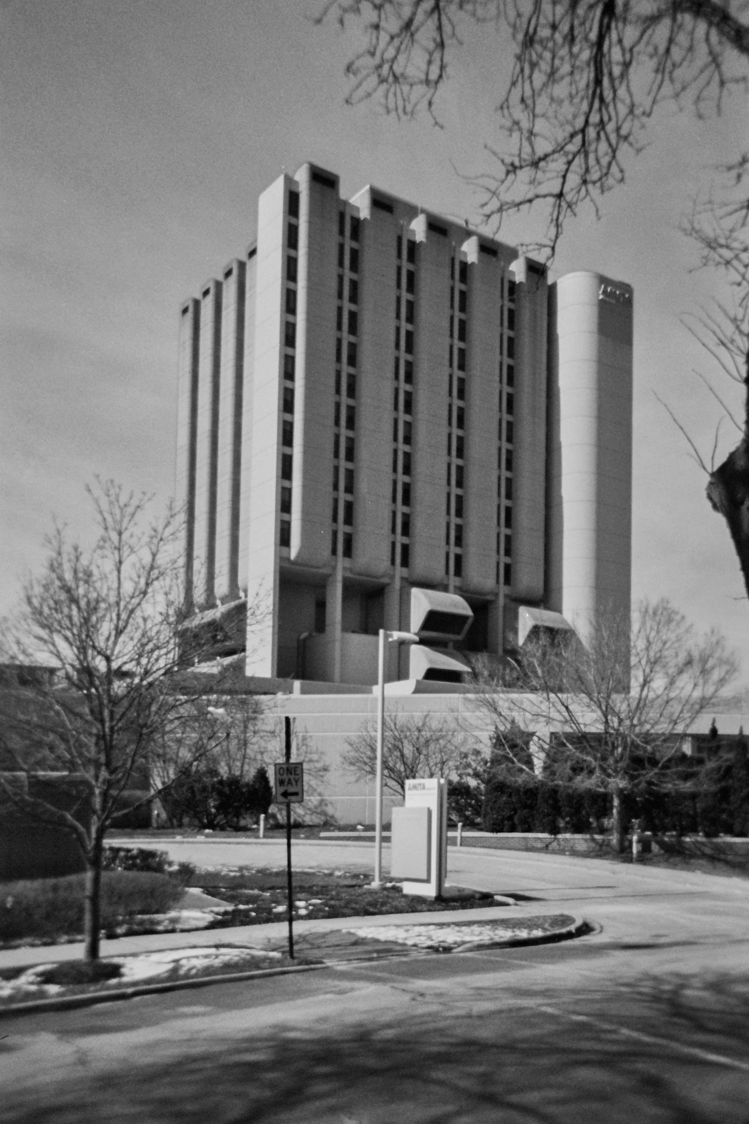 Black and white photo of a tall, modern building with vertical lines and cylindrical sections, surrounded by leafless trees and a street with traffic signs.