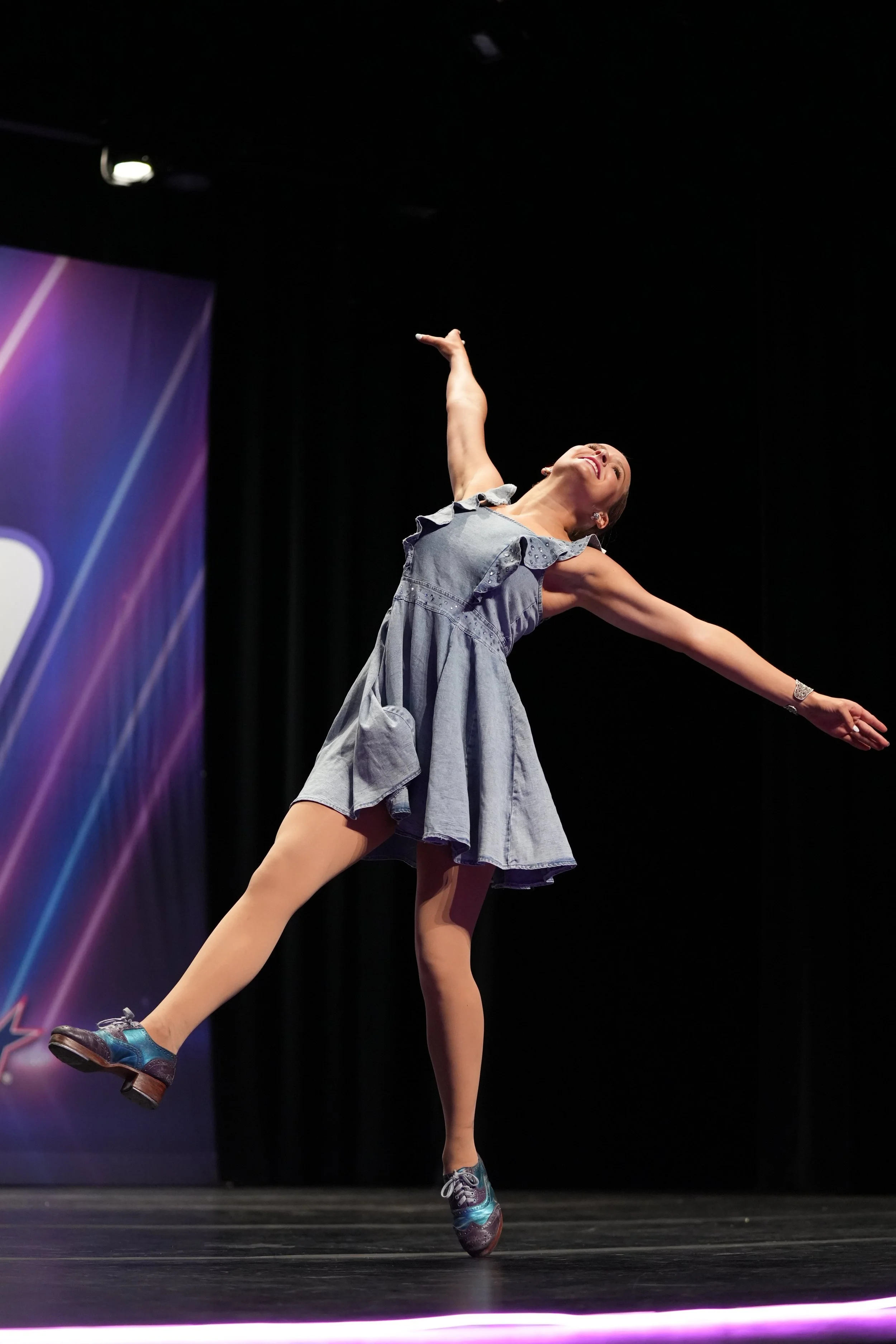 A dancer performing on stage in a denim dress with arms outstretched and one leg raised.