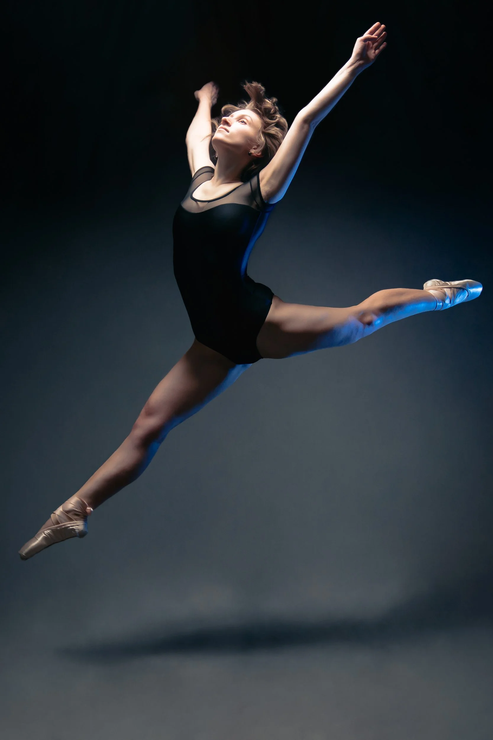 Ballet dancer in black leotard performing a leap with arms extended upward on a dark background.