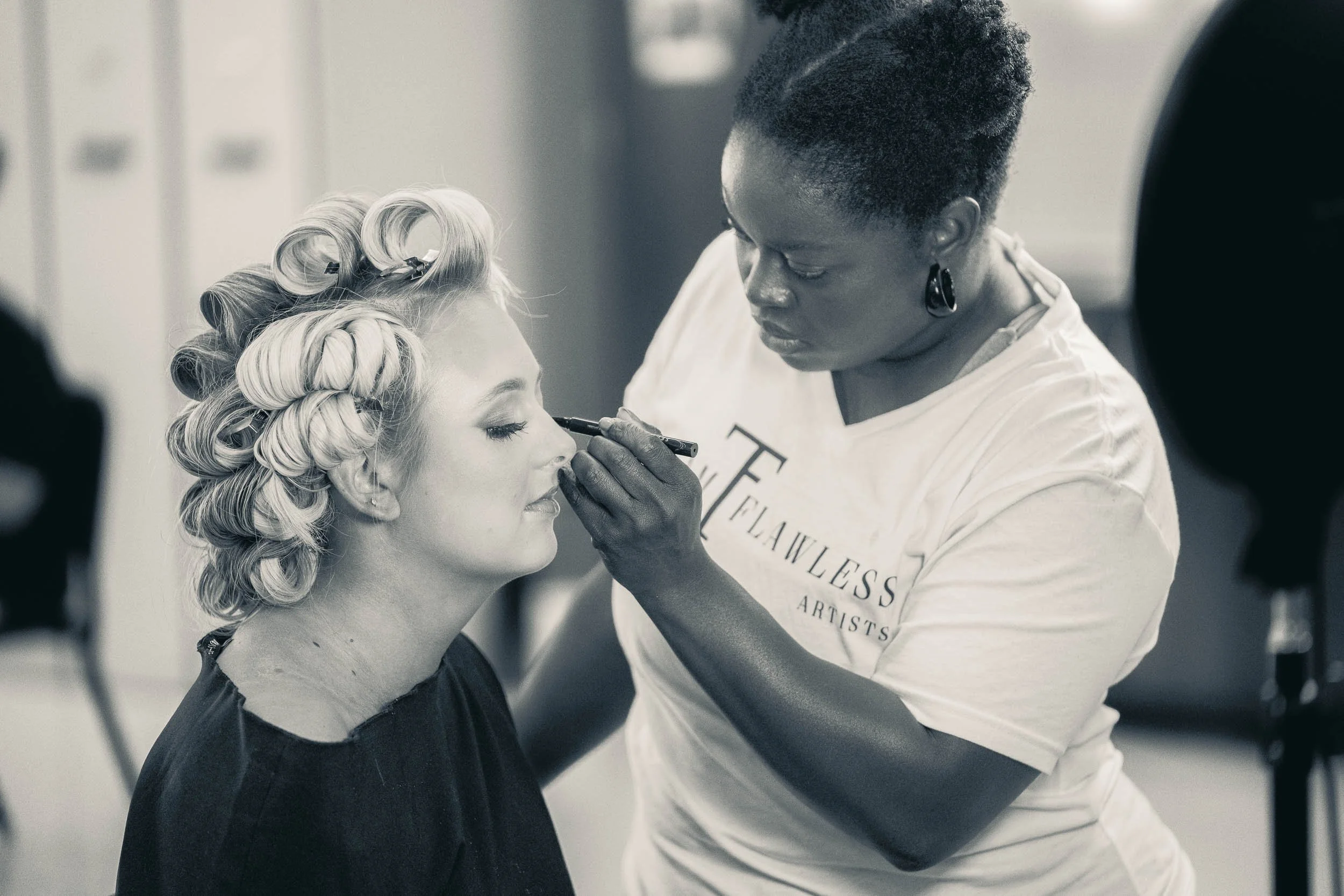 Makeup artist applying makeup on a woman's face with curlers in her blonde hair as she sits patiently.