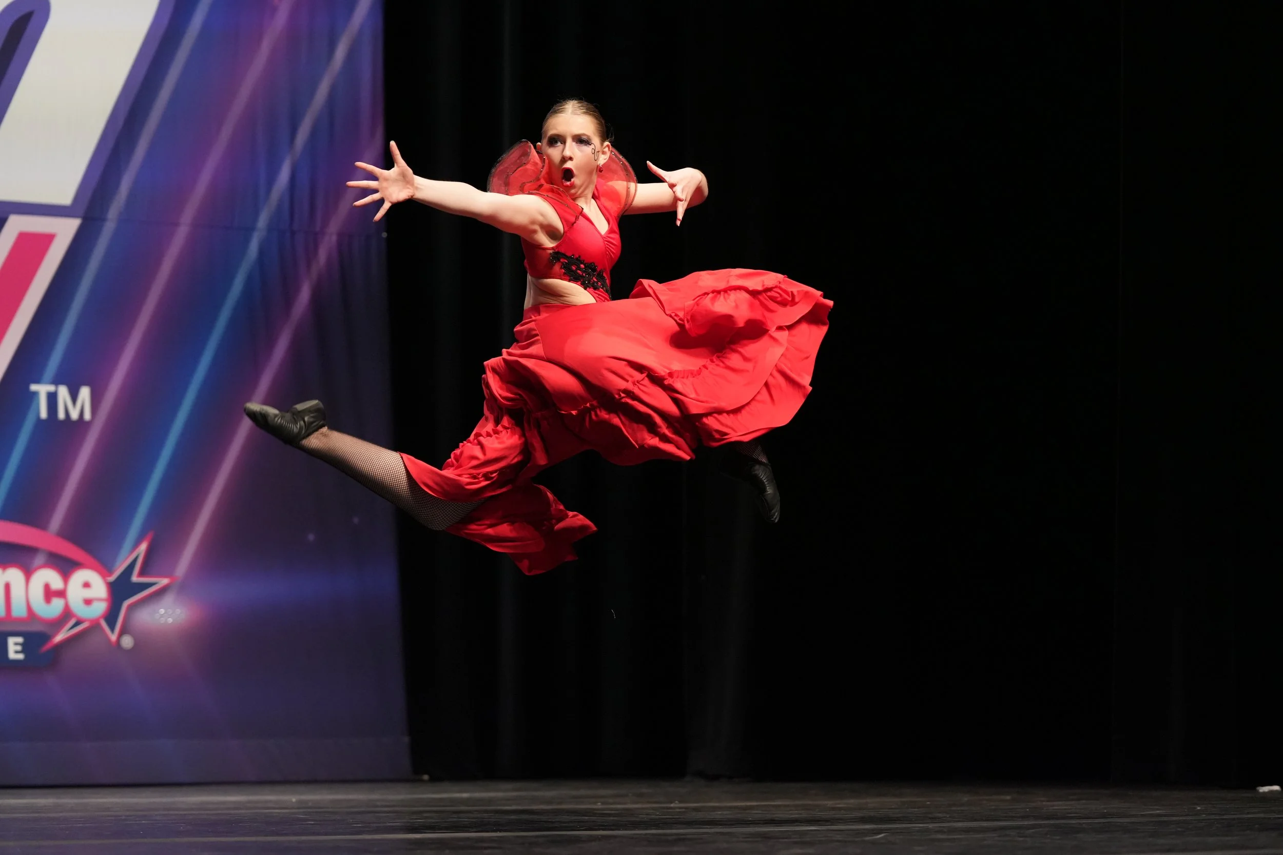 A female dancer in a red dress performing a mid-air jump on stage at a dance competition.