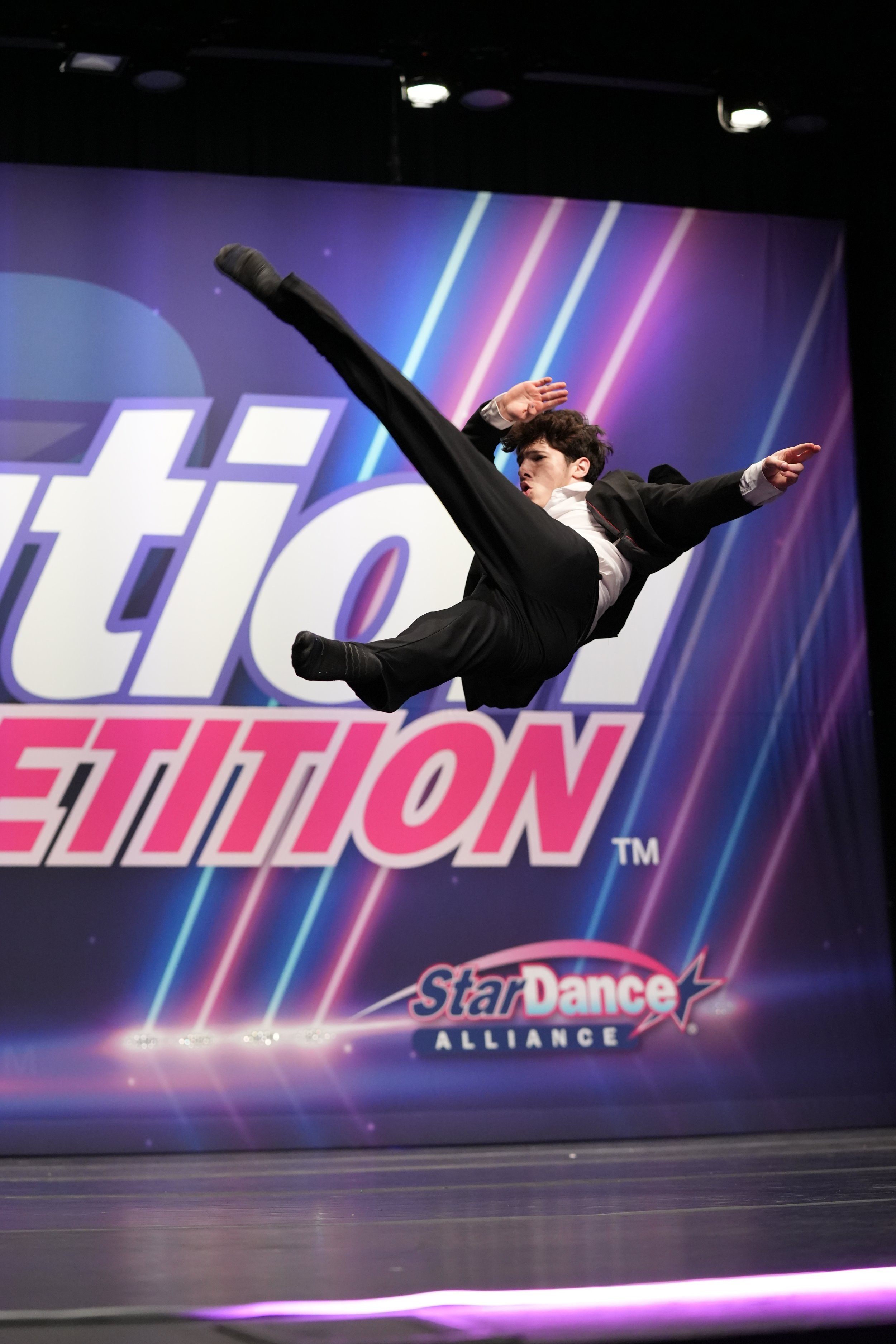 A male dancer in a suit performing a leap on stage with a colorful 'StarDance Competition' backdrop.