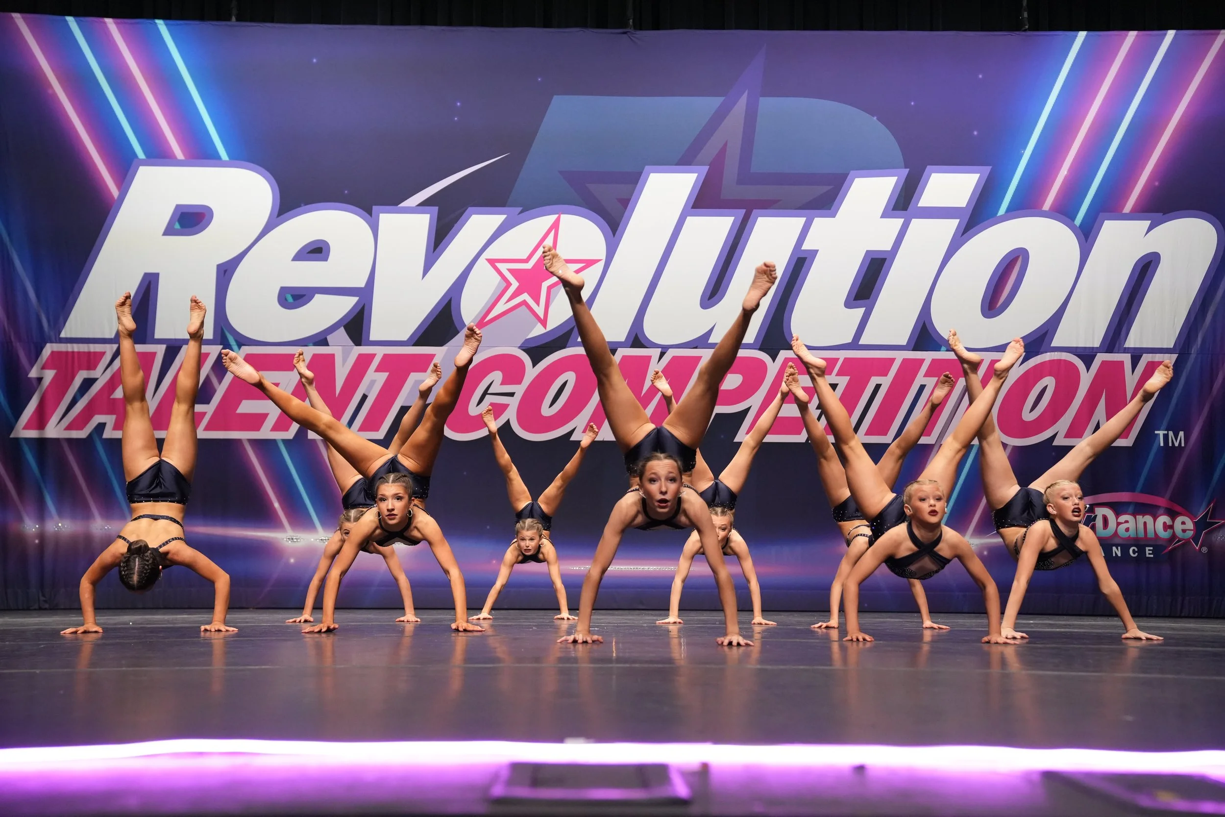 A group of young girls in dark dance costumes performing a handstand and split on stage during a dance competition with a colorful backdrop that says 'Revolution Talent Competition'.