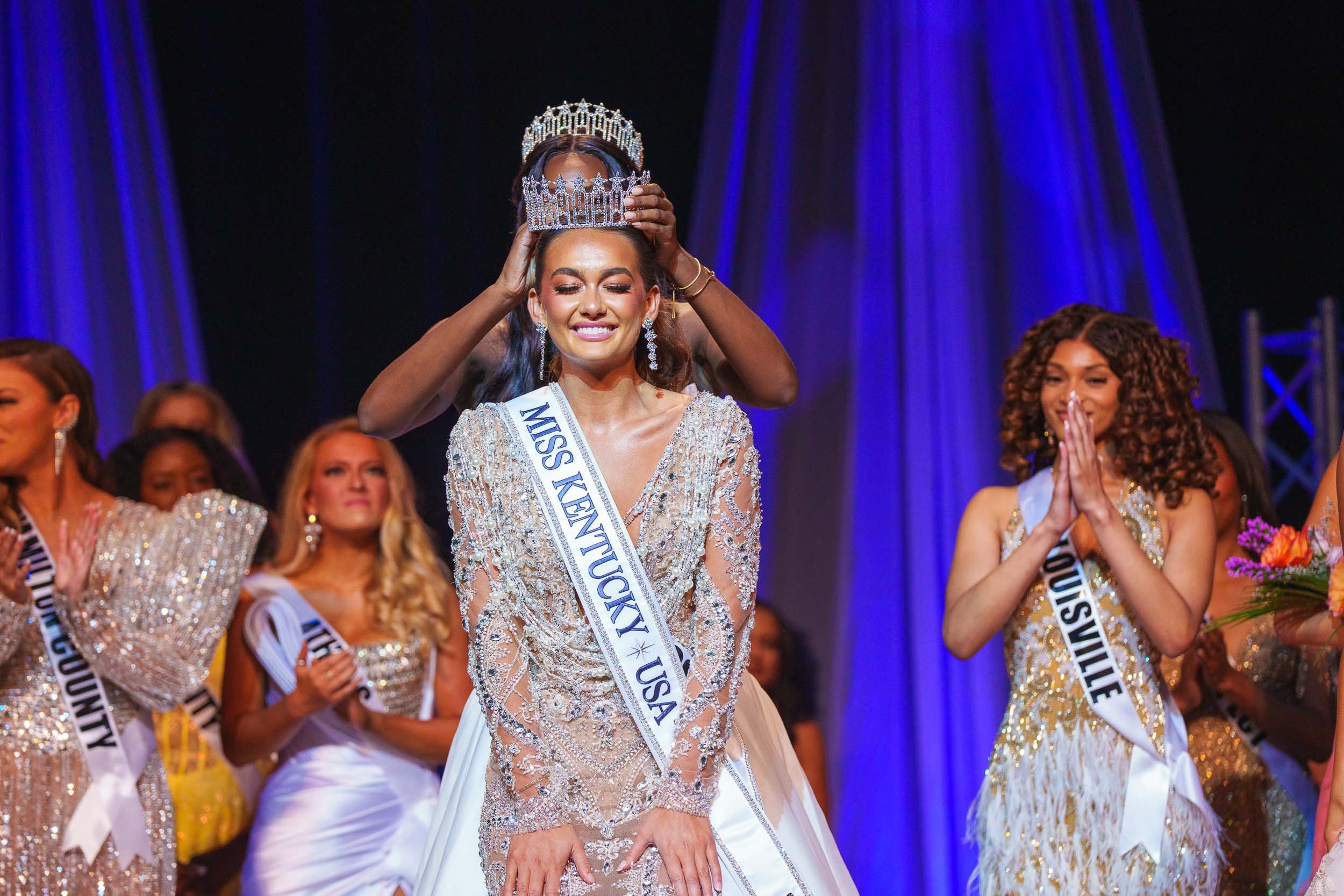 Miss Kentucky 2025 being crowned, smiling on stage with other contestants clapping around her.