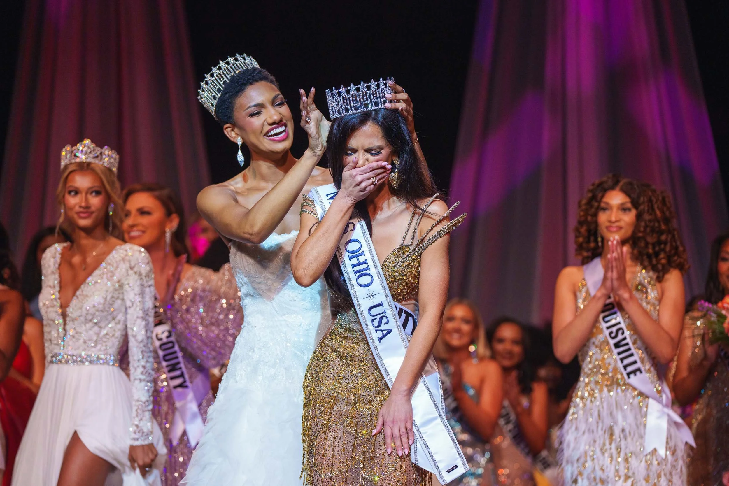 Miss Ohio 2025 winner crowned on stage, woman placing crown on another woman, others clapping and smiling, colorful stage curtains in background.