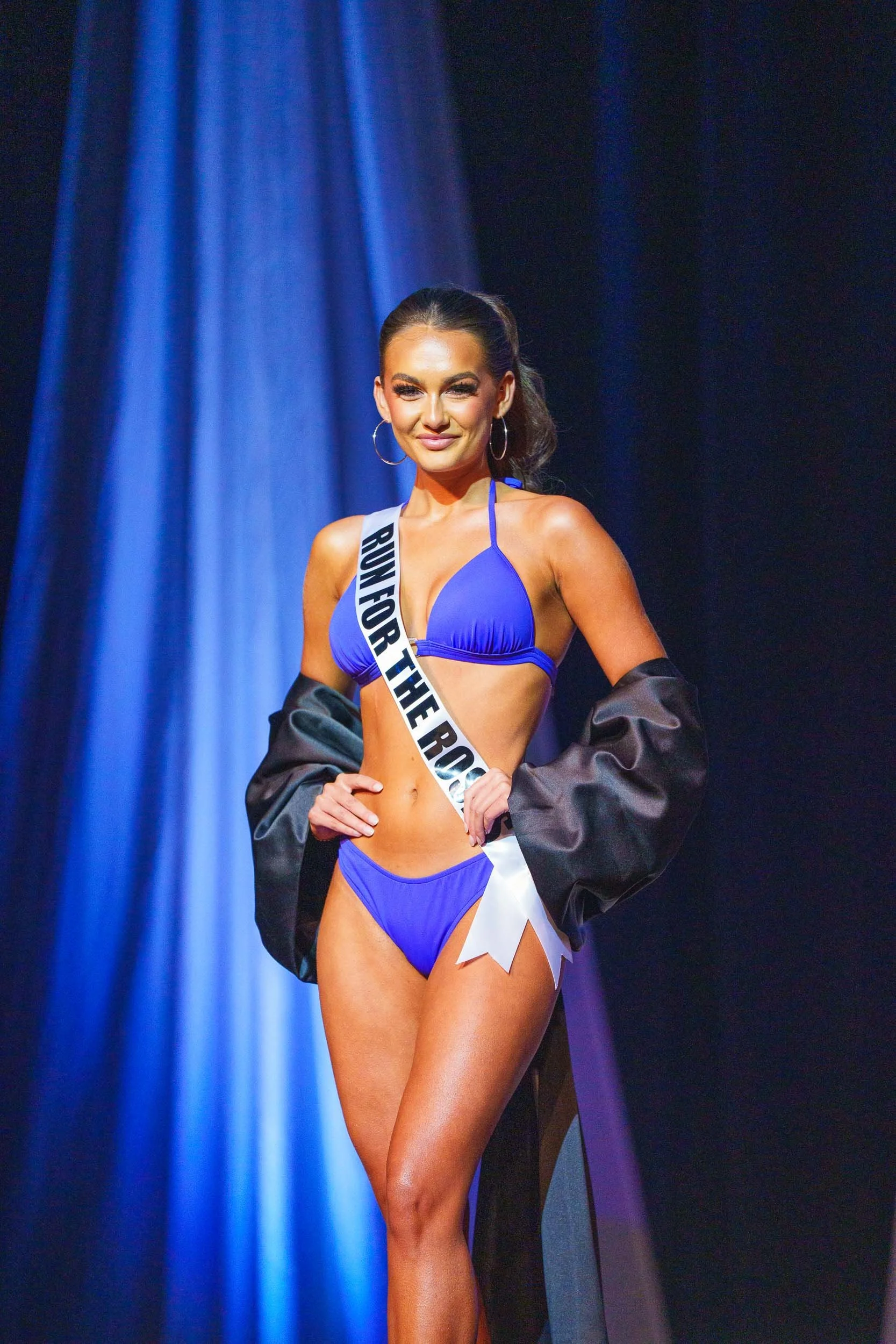 A woman in a blue bikini and large hoop earrings on stage wearing a sash that reads "RUN FOR THE R" and a black jacket draped over her arms, standing in front of a blue curtain.