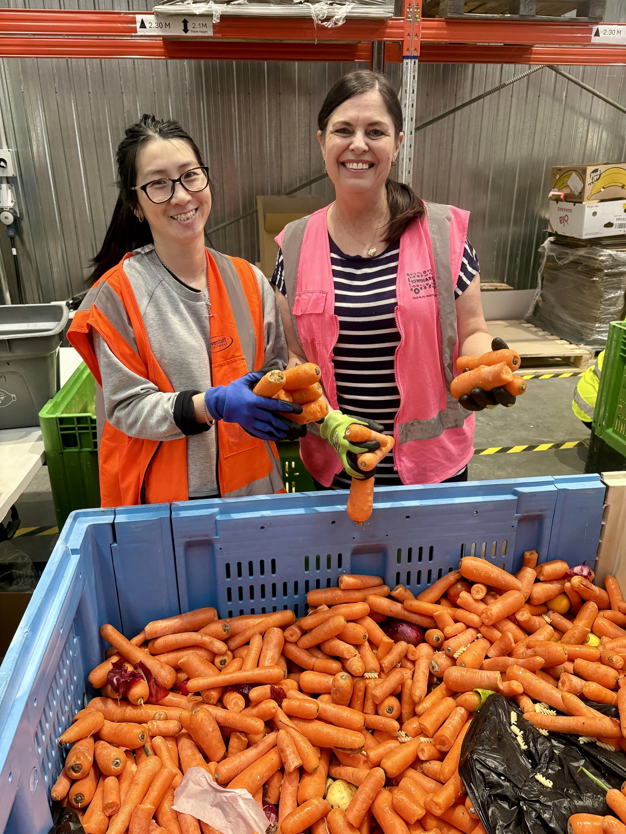 Two women smiling and holding carrots in a warehouse or market setting with stacked crates and shelving.