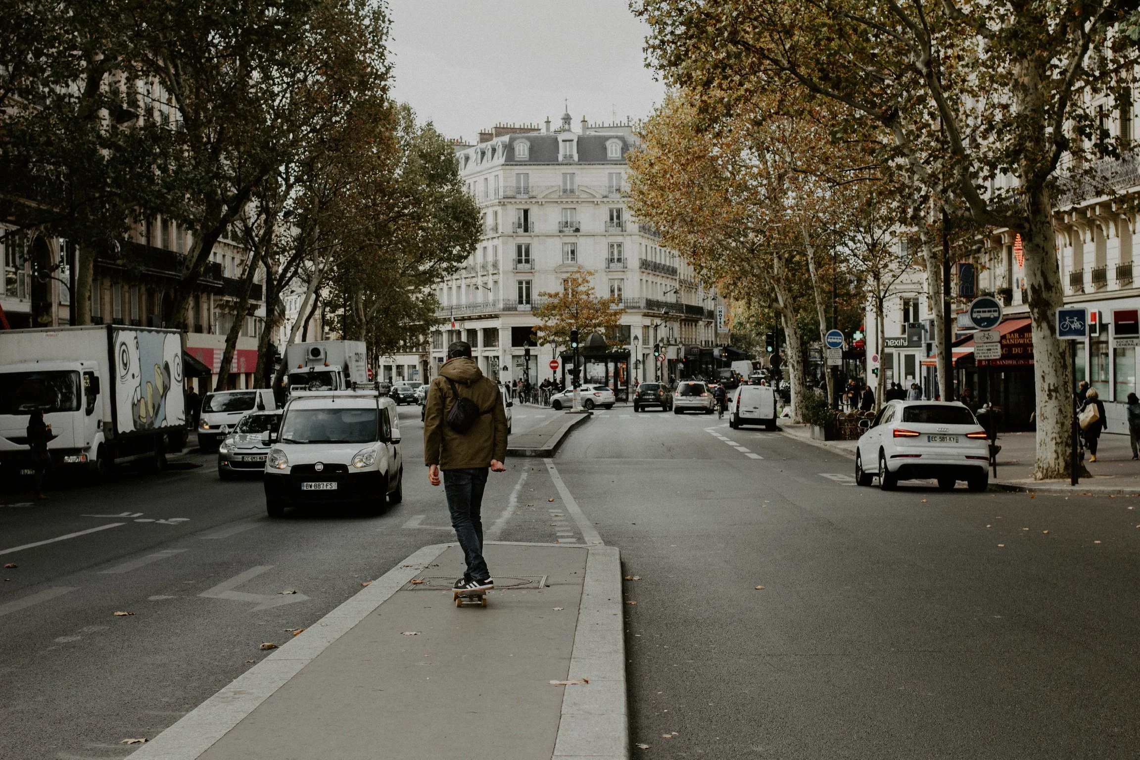 A person skateboarding on a city sidewalk with trees, cars, and buildings around, in a European city, on a cloudy day.