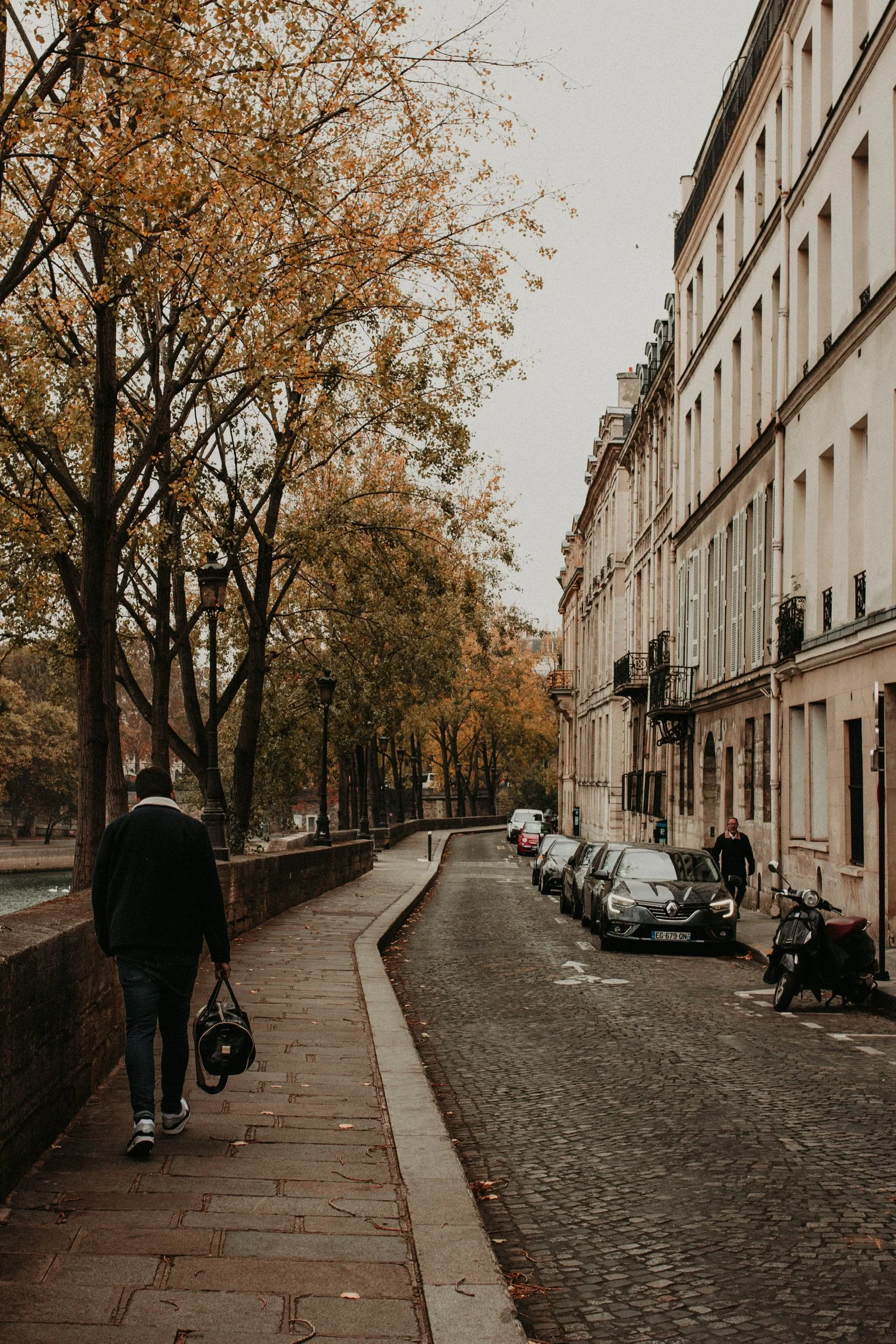 A man with a backpack walking on a cobblestone sidewalk next to parked cars and a row of white multi-story buildings with balconies in a city street with autumn trees.