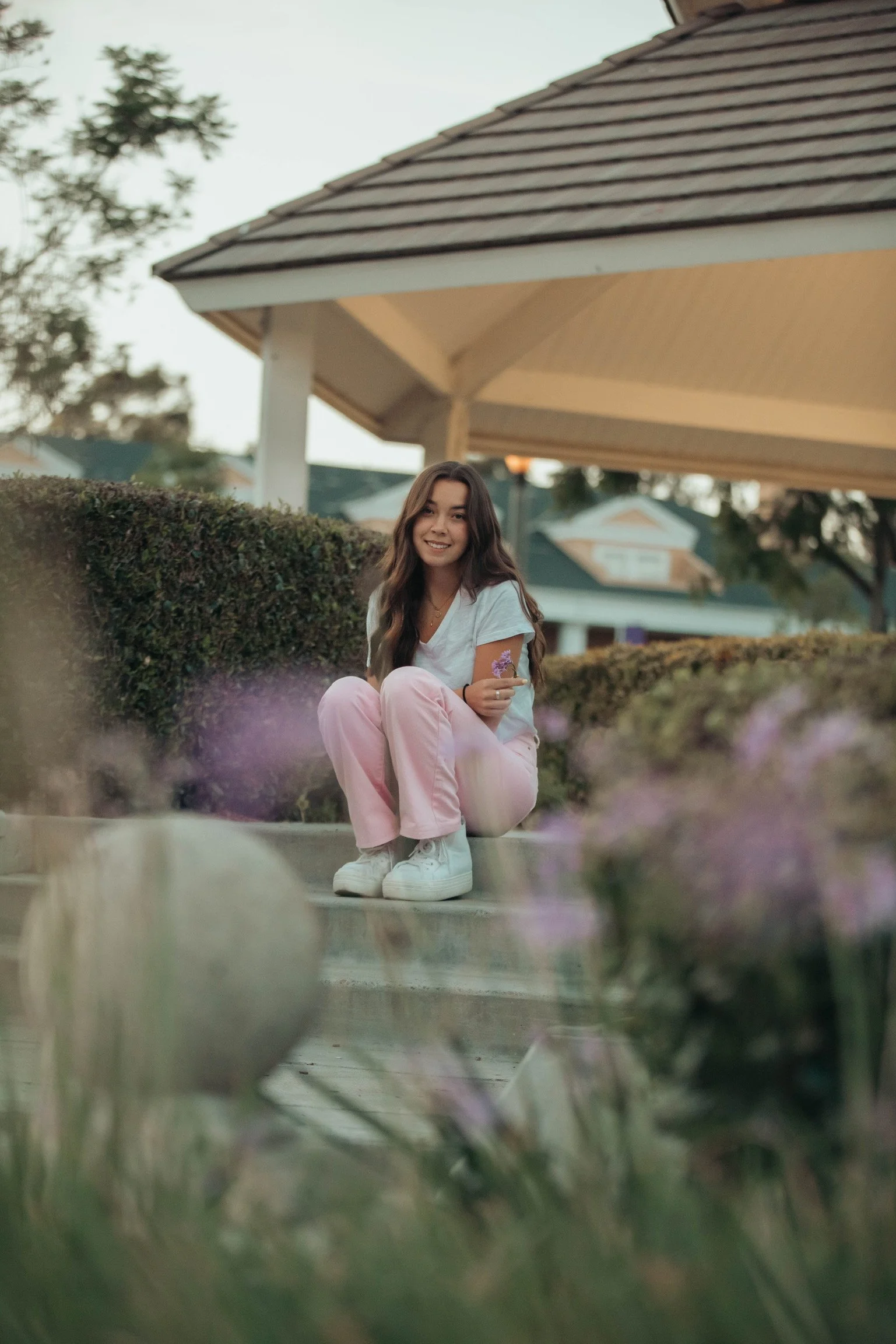 A young woman with long wavy brown hair sitting on the steps outside a house, surrounded by bushes and flowers, smiling and looking at the camera.