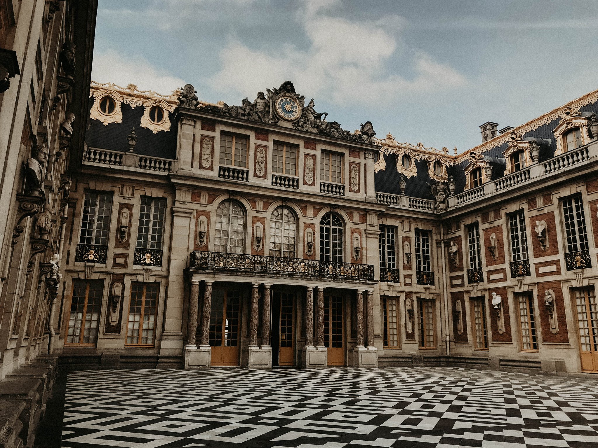 Facade of a grand historic building with ornate architecture, multiple windows, balconies, decorative sculptures, and a checkered black-and-white floor courtyard.