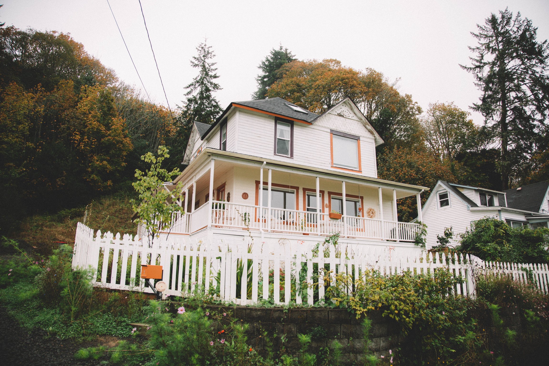 A multi-story white house with a front porch and railing, surrounded by a white picket fence and lush greenery, trees, and autumn foliage in the background.