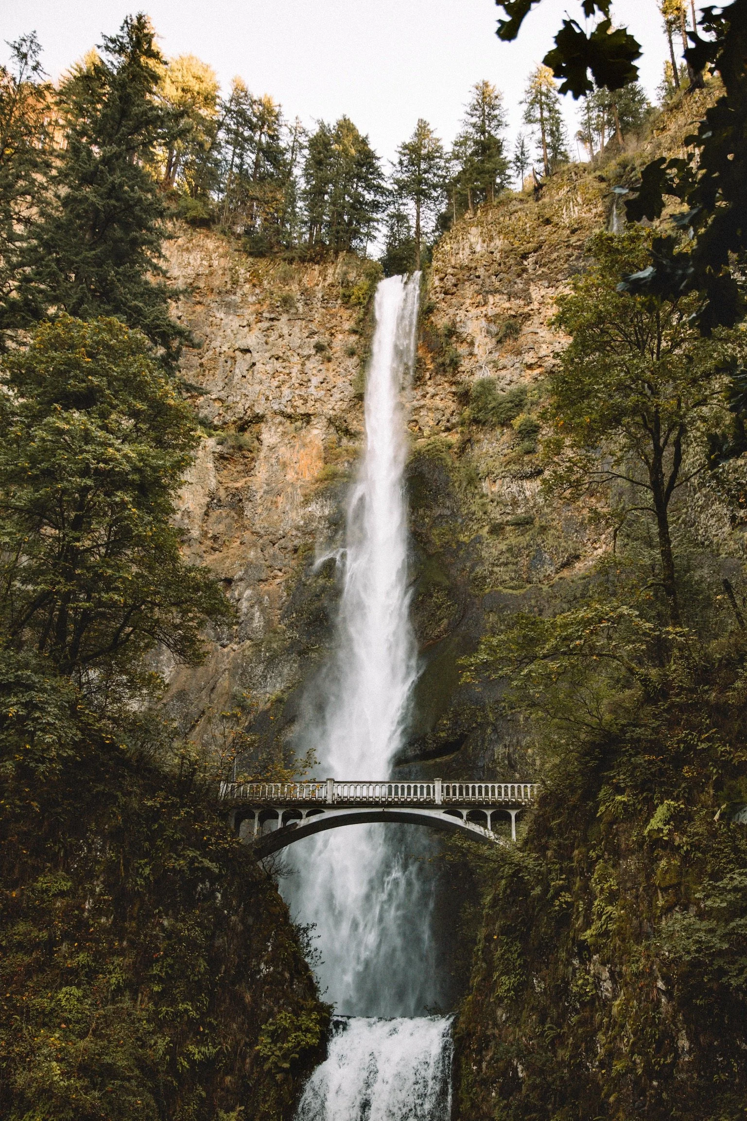 Tall waterfall flowing down a rocky cliff surrounded by green trees, with a bridge crossing in the middle of the waterfall.