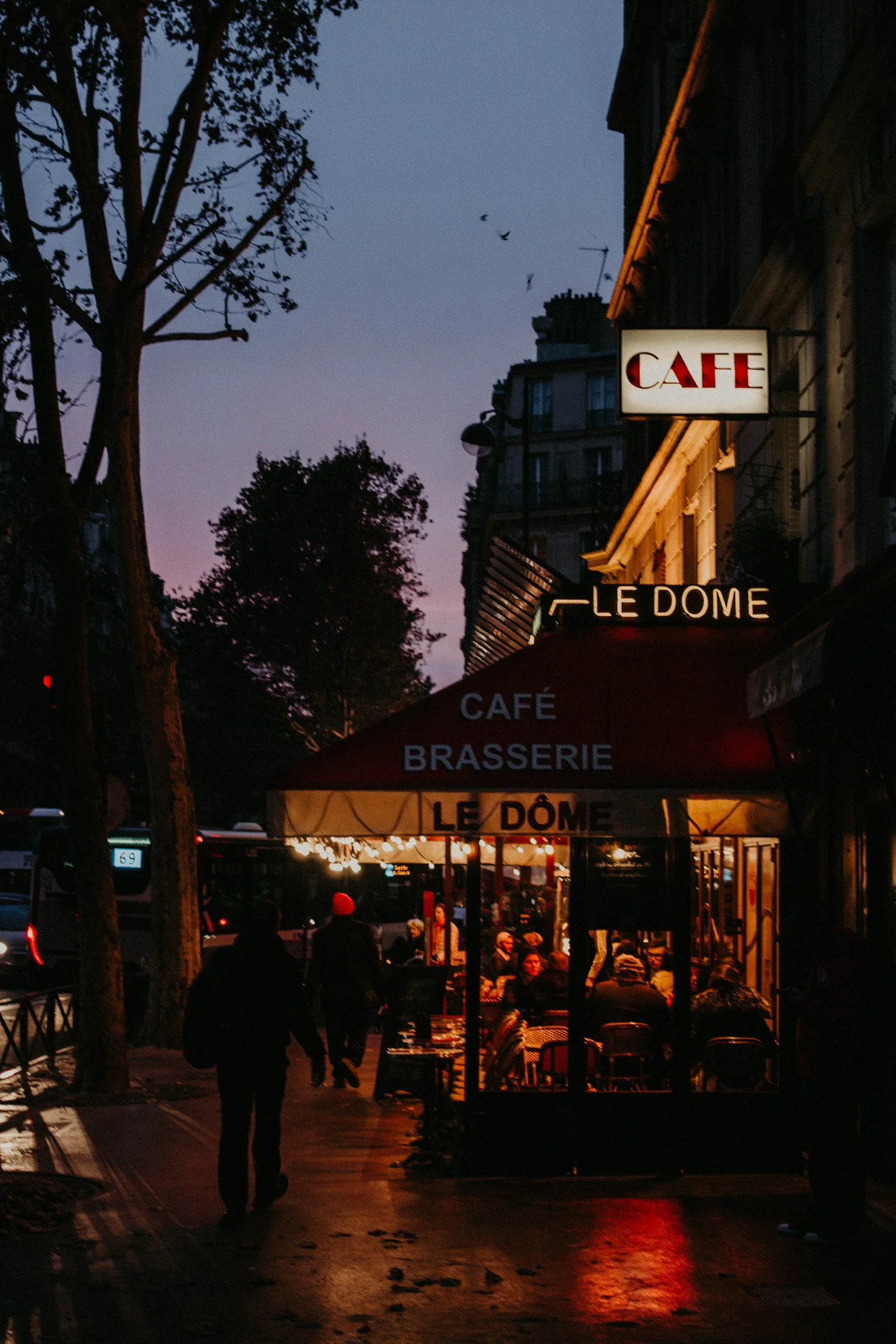 A Parisian street scene at dusk with a restaurant called 'Le Dôme' featuring outdoor seating illuminated by warm lights, people walking on the sidewalk, and signs for 'Café' and 'Brasserie' above the establishment, with the sky transitioning from twi
