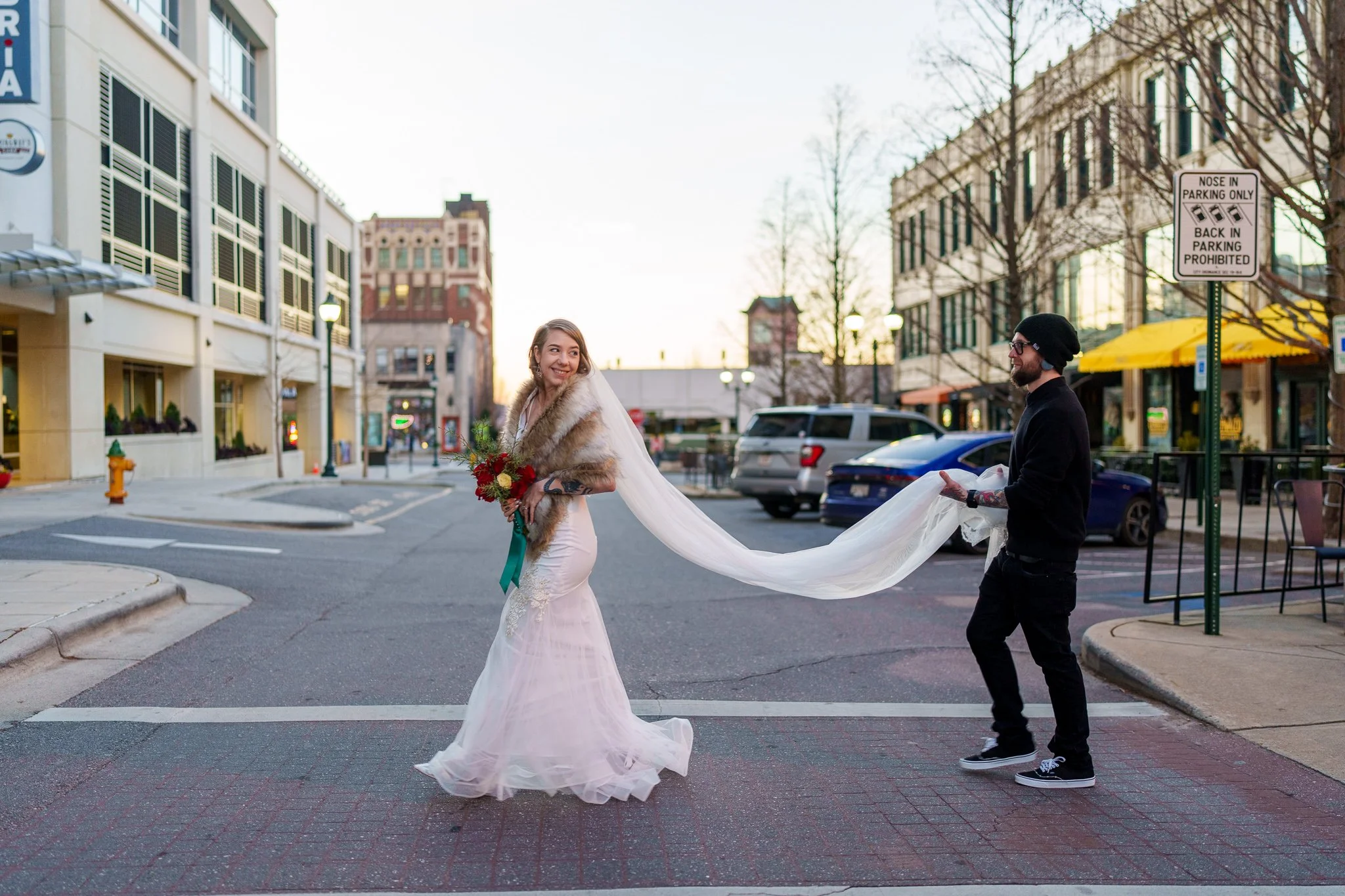 A bride in a white wedding dress and fur stole holding a bouquet of flowers is smiling for a photo in the street while a behind-the-scenes partner in dark clothes holds her long train.