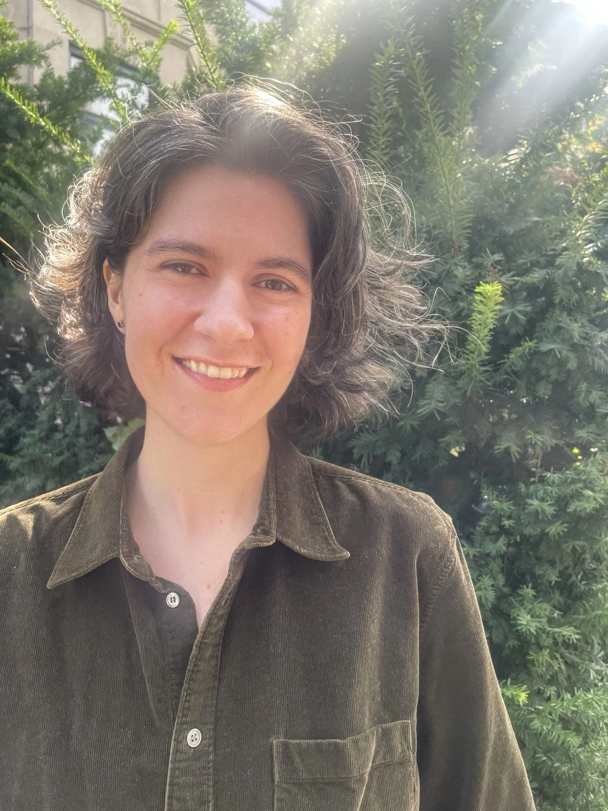 Jackie stands outside in the sunshine in front of greenery with chin-length brown and grey wavy hair, wearing a green corduroy button-up.