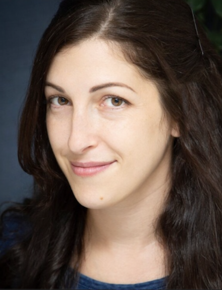 Headshot of a white woman with brown hair and brown eyes, wearing a blue cotton shirt, looking into the camera and smiling softly against a blurred background. 