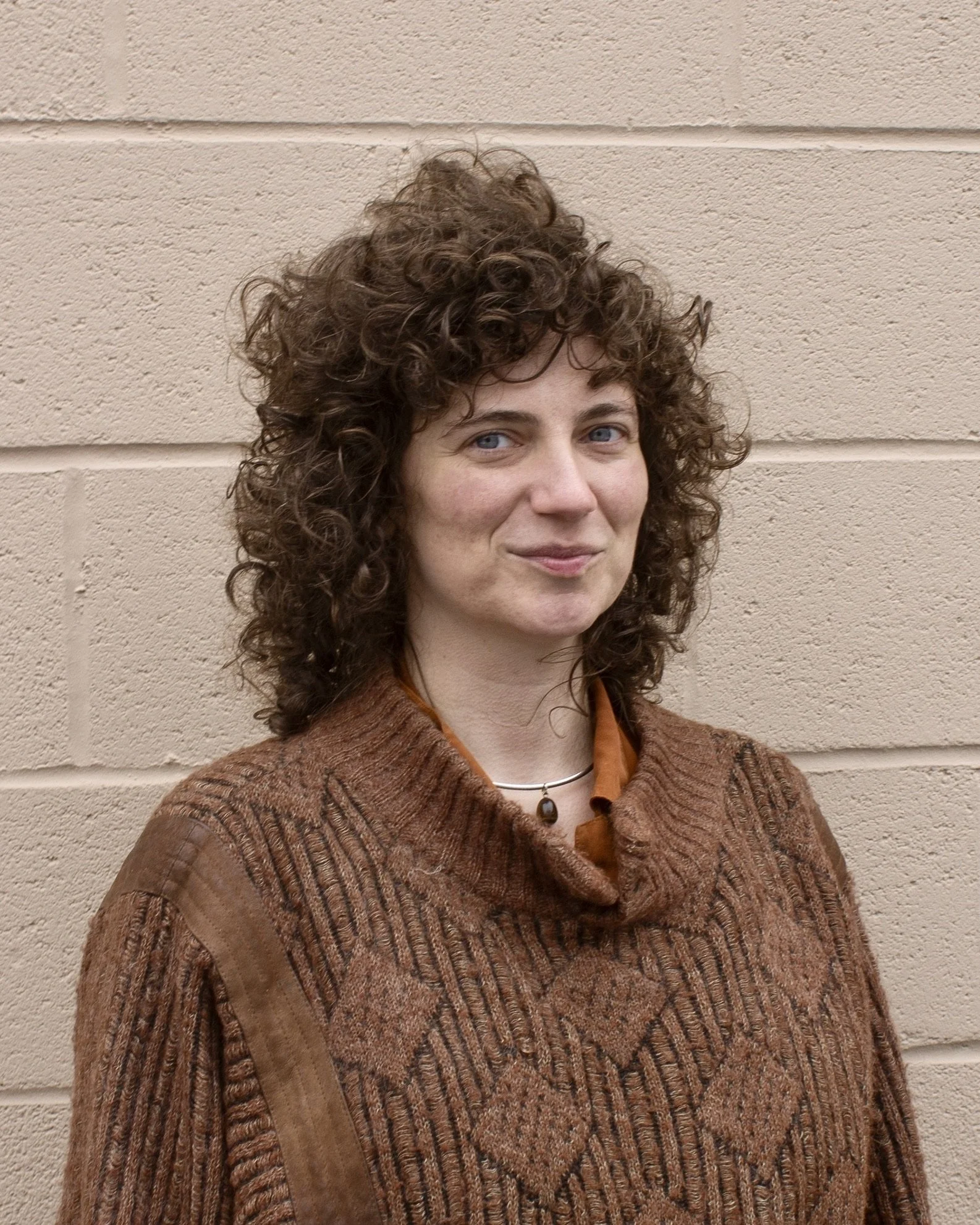 A curly-haired, blue-eyed white femme wearing an oversized brown sweater stands against a tan concrete wall, meeting the camera with a grounded gaze and warm smile.
