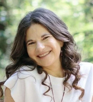 A warm, smiling woman with long, wavy brown hair tilts her head kindly in an outdoor setting. She wears a white top and a delicate necklace, projecting a friendly, trustworthy presence. 