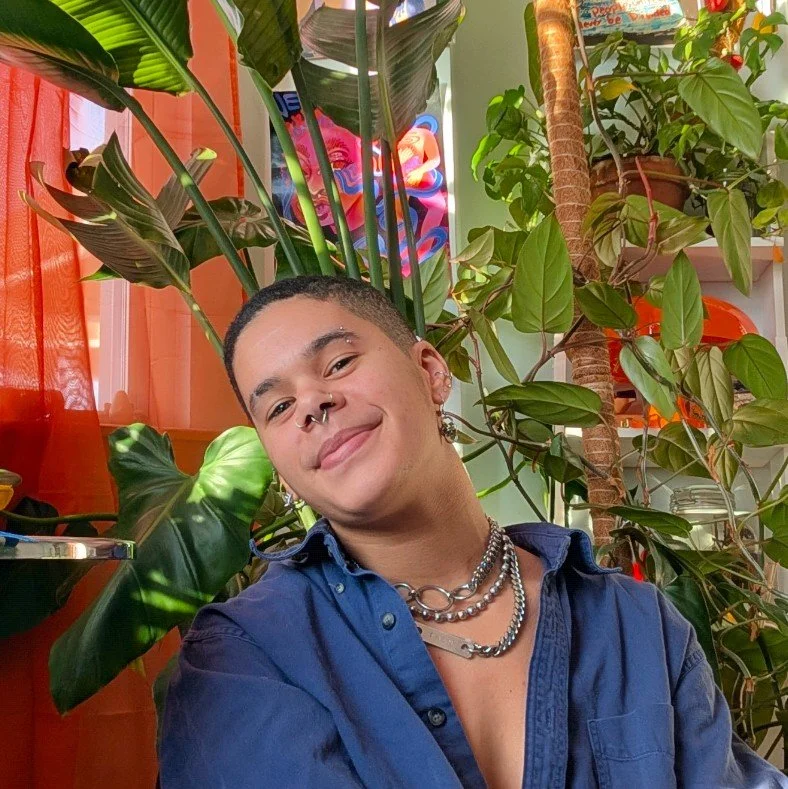 A light-skinned black agender person with short black hair is sitting indoors in front of their plants. They are wearing a blue shirt and smiling at the camera with a slight tilt in their head.