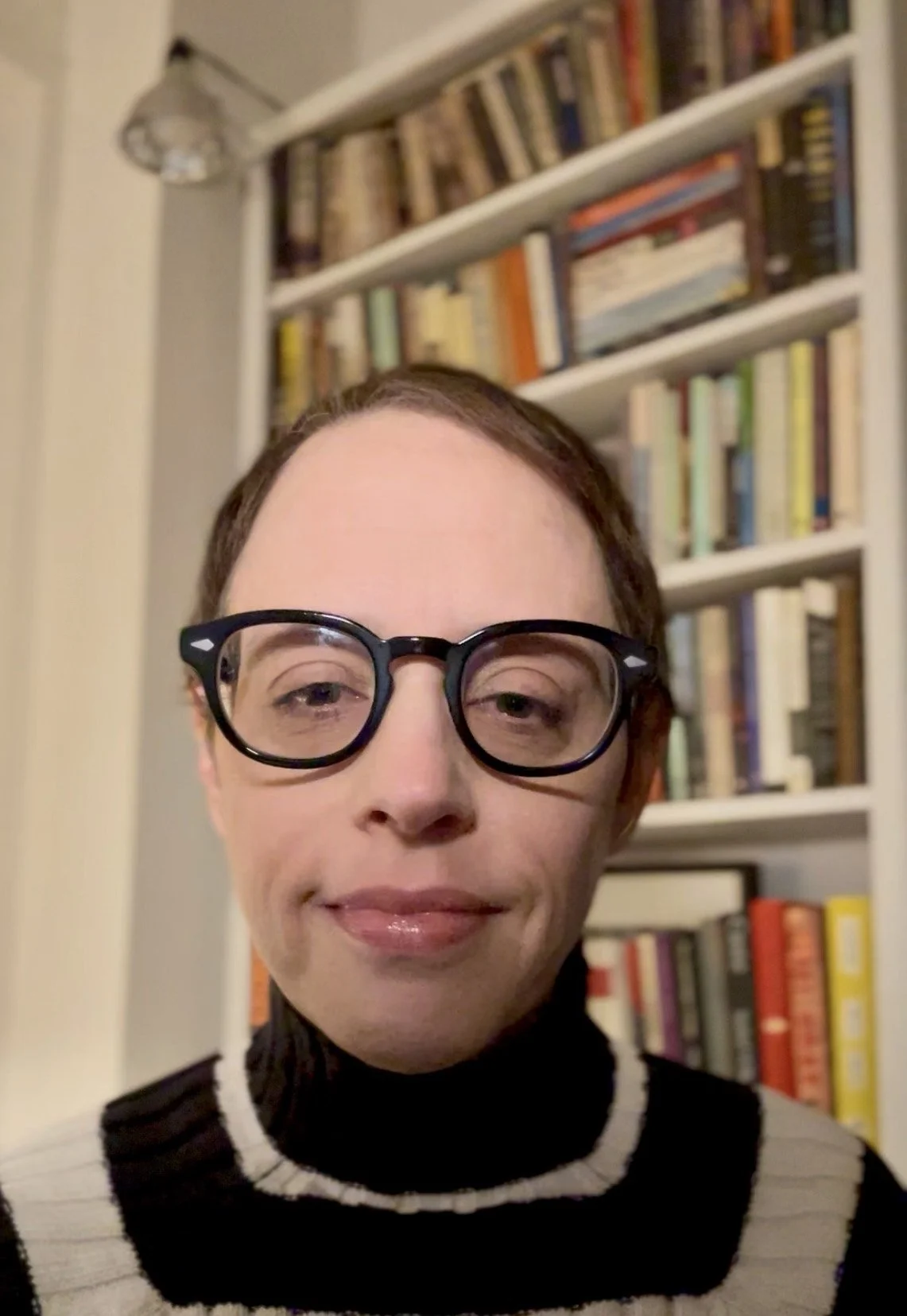Portrait of Abbie Ruth, a white woman with short brown hair, wearing glasses and a black-and-white sweater, smiling at the camera, with a bookshelf visible in the background.