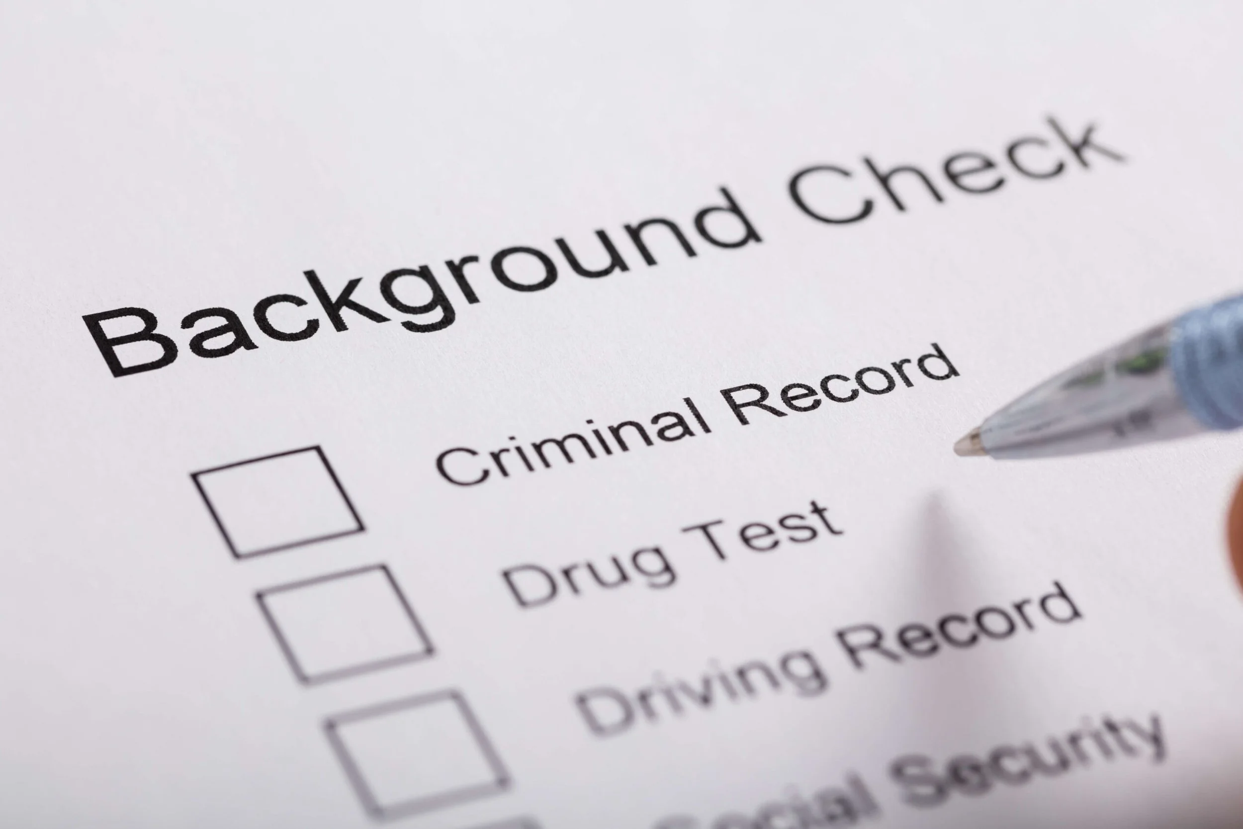 Close-up of a paper titled 'Background Check' with checkboxes for Criminal Record, Drug Test, Driving Record, and Digital Security, with a pen pointing at the paper.