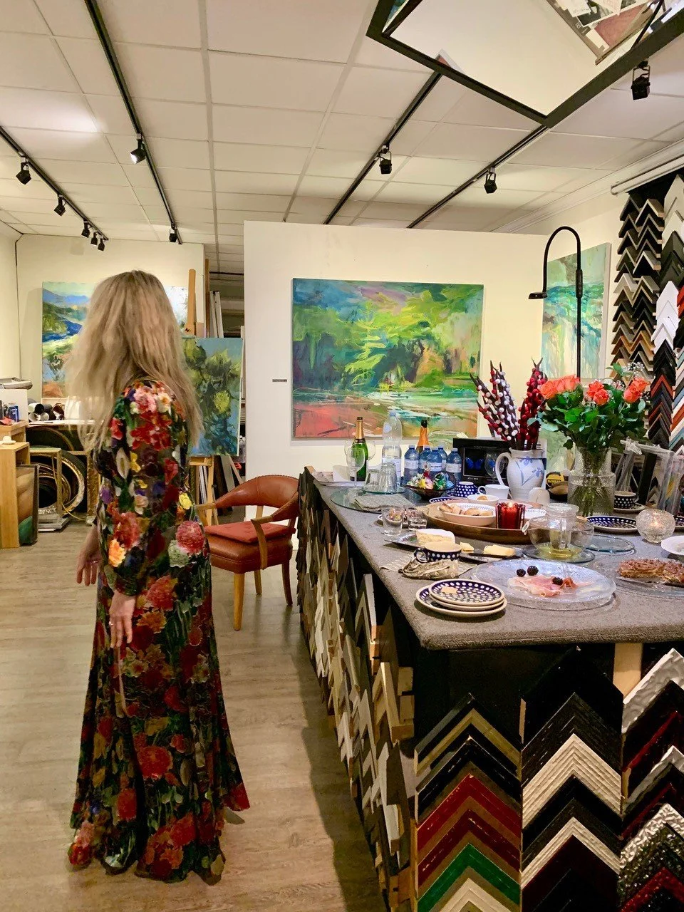 Interior of an art studio with a woman in a long floral dress, standing near a table filled with snacks, drinks, and decorative items. The background features colorful landscape paintings on white walls, and sample frame corners arranged on the wall.