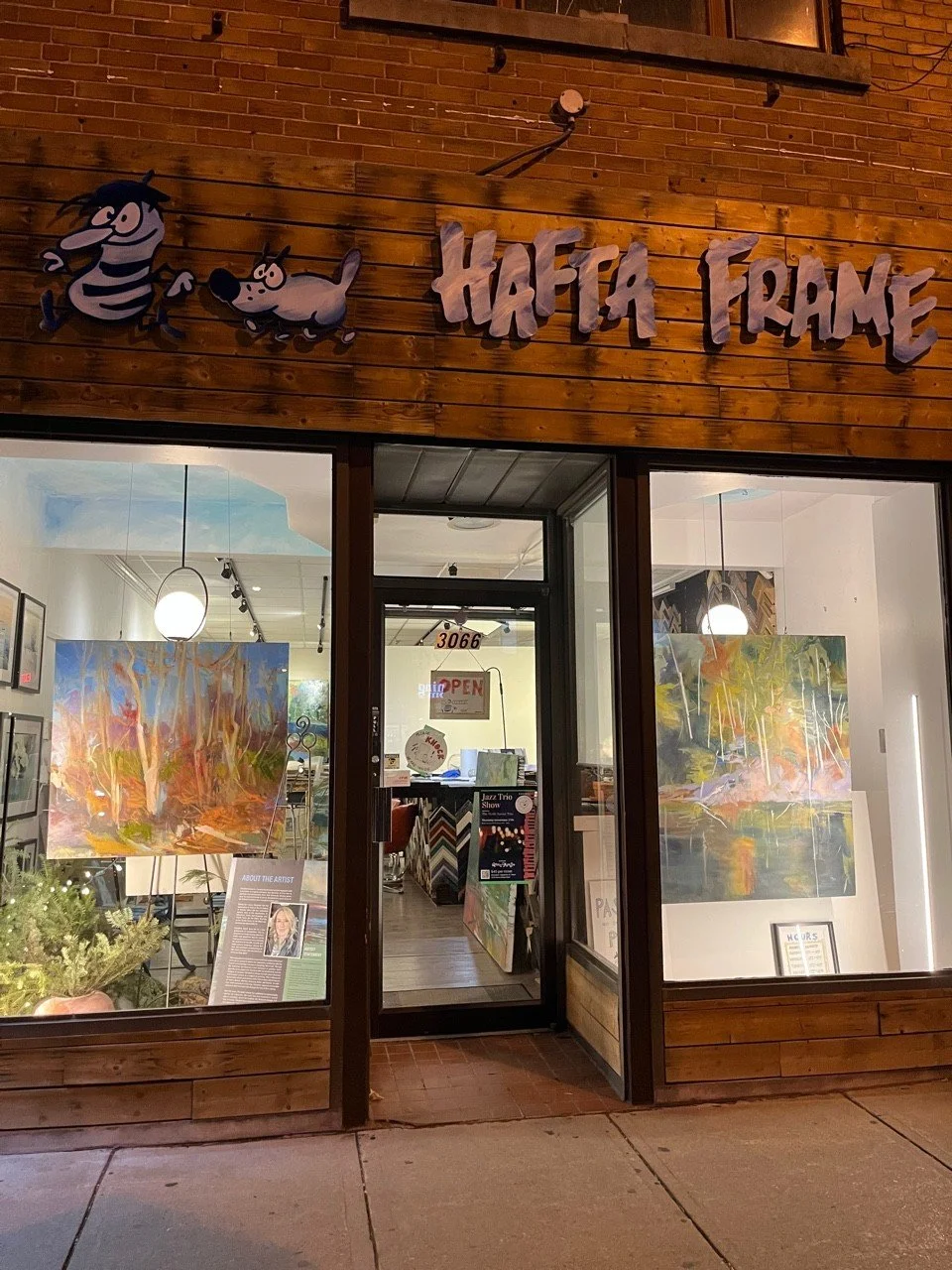 The storefront of Hafta Frame art gallery features a wooden exterior with animated characters and the name in stylized letters. Inside, there are colorful paintings on display, with lighting and an informational sign about the artist visible through the large windows.