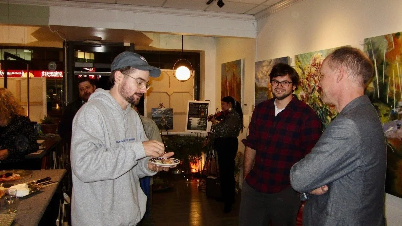 People talking and socializing in an art gallery or cafe with paintings on the wall, some enjoying food, and a person playing a violin in the background.