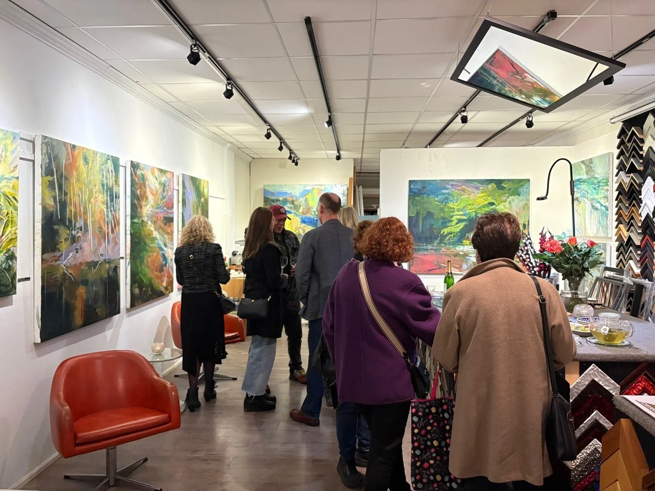 People attending an art gallery, viewing colorful abstract paintings on the walls, with some engaging in conversation, and a table with flowers and framed artworks in the foreground.