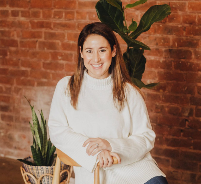  sitting woman pose for a photo in front of a brick wall. The woman is smiling and wearing a cream colored sweater. There are live plants in the background