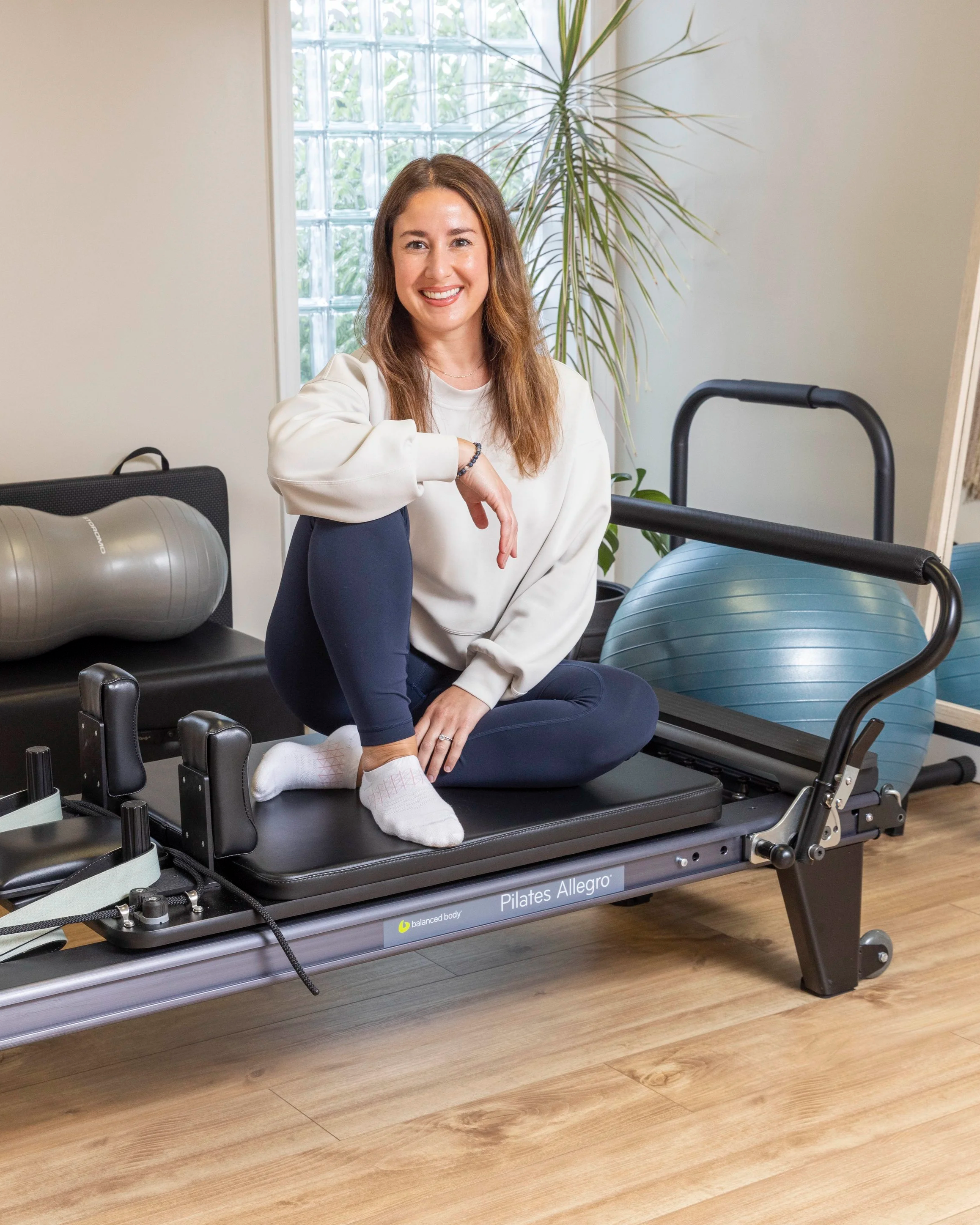 A woman sitting cross-legged on a Pilates reformer machine in a well-lit room with a large window and a plant in the background. She is smiling and dressed in a white sweatshirt and navy pants.