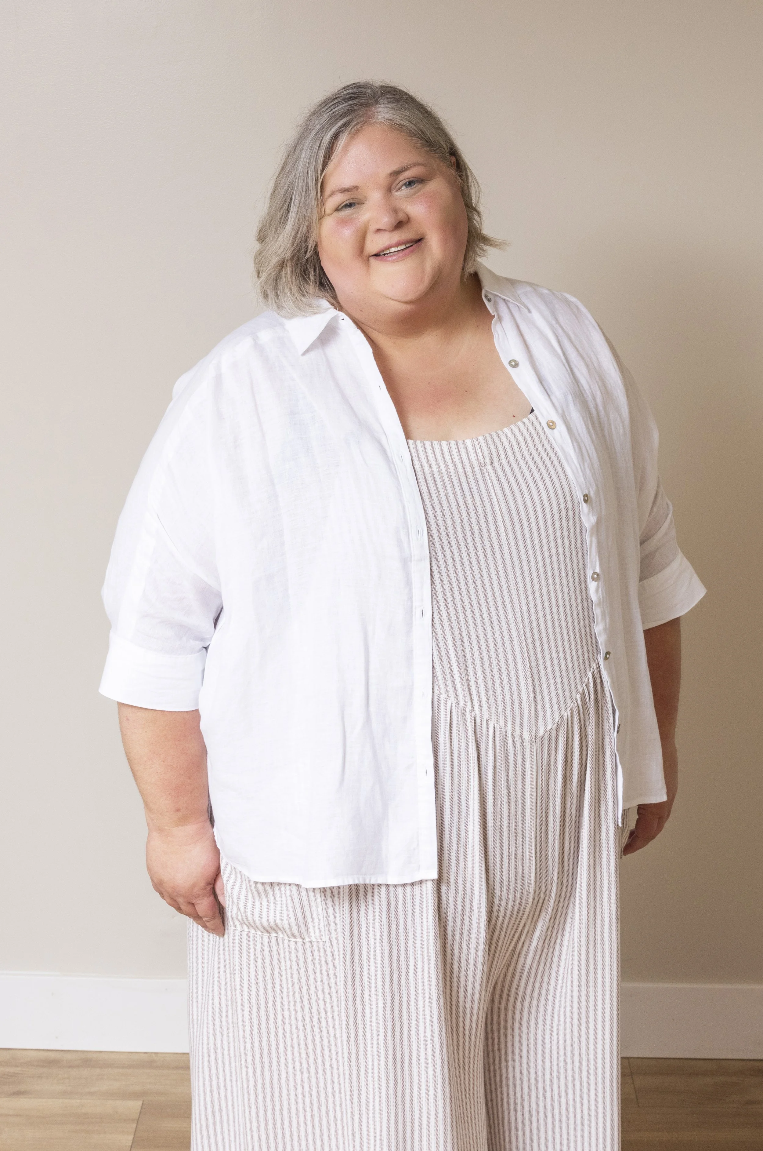 A grey-blond woman wearing a beige and white striped romper and white button down open in front smiles at the camera
