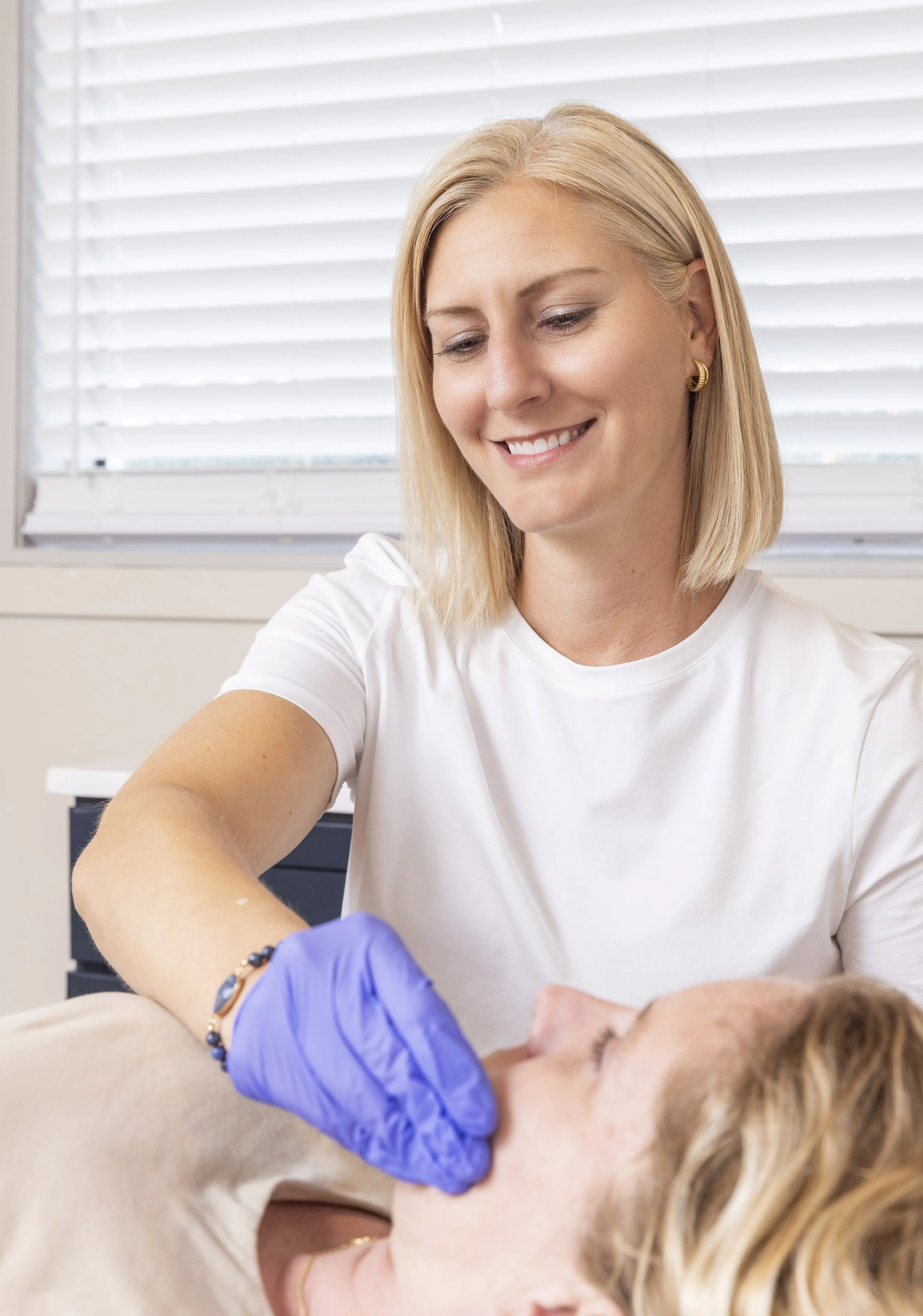 A female physical therapist with blonde hair and gold hoop earrings smiling while providing intraoral treatment for TMJ to a client lying on a treatment table, with a background of window blinds.