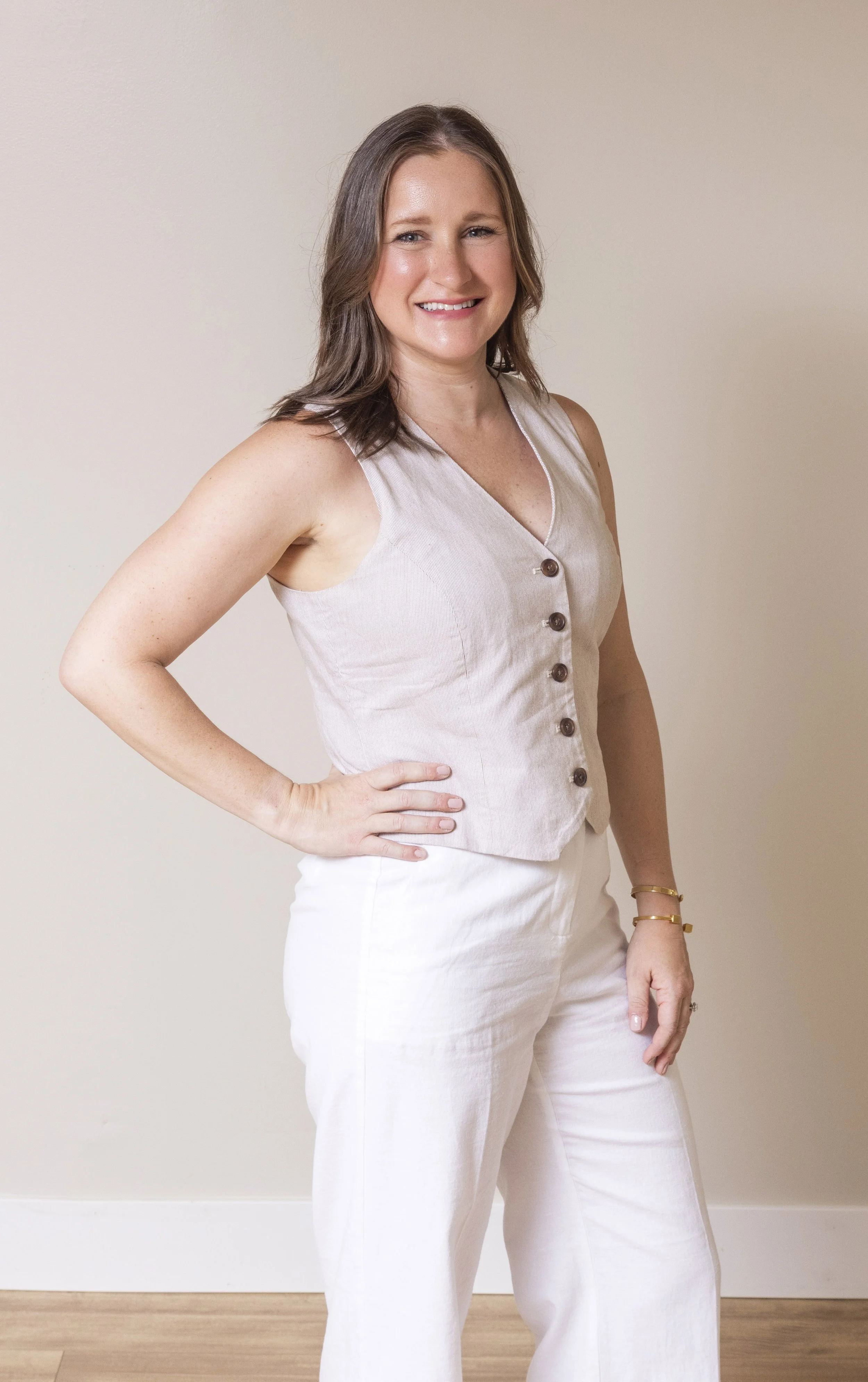A woman with shoulder-length brown hair, smiling, wearing a beige sleeveless top with buttons down the front, and white pants, standing indoors against a plain light-colored wall.
