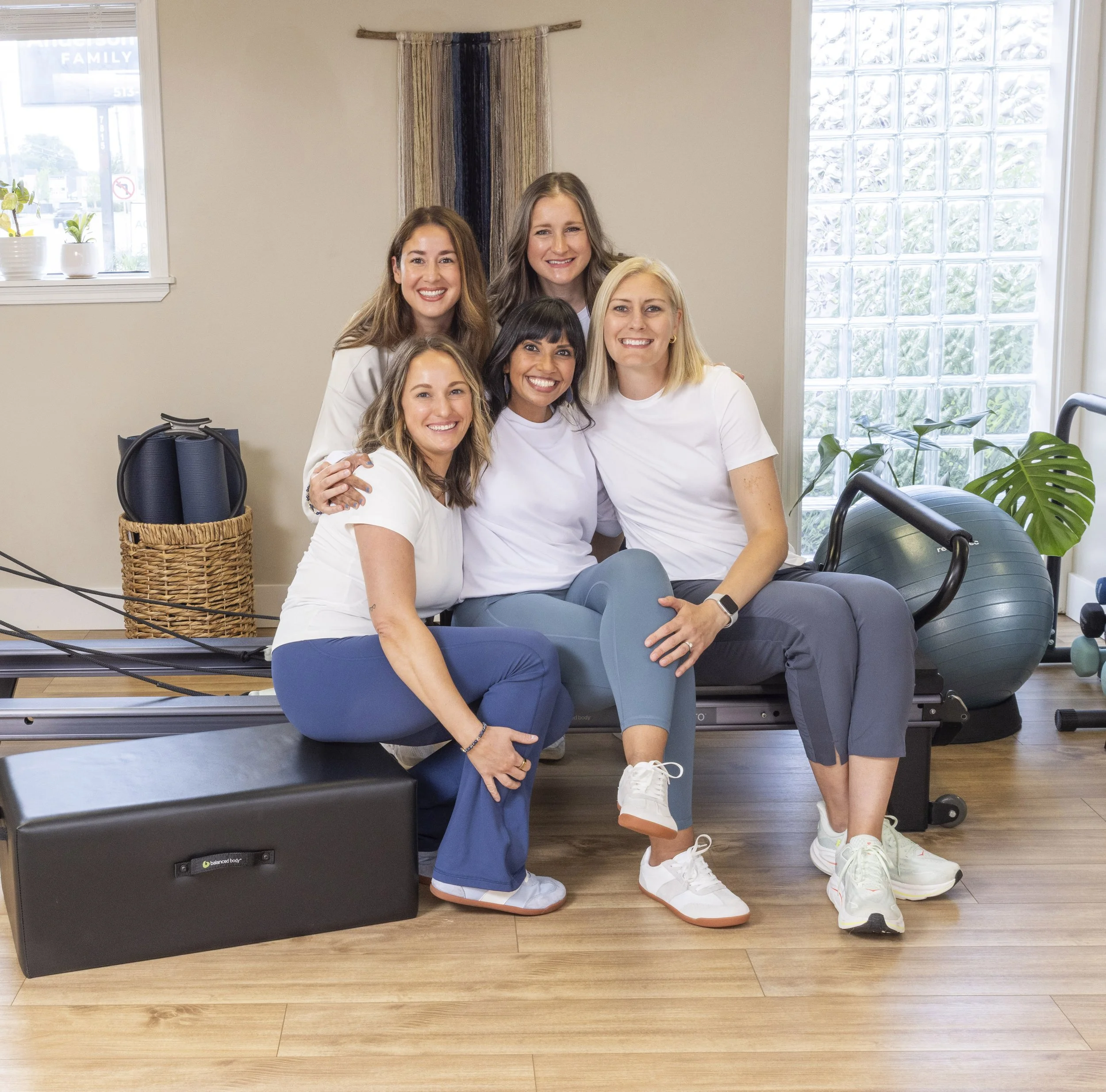 Group of six women in a fitness studio, smiling and sitting together on exercise equipment surrounded by natural light and houseplants.