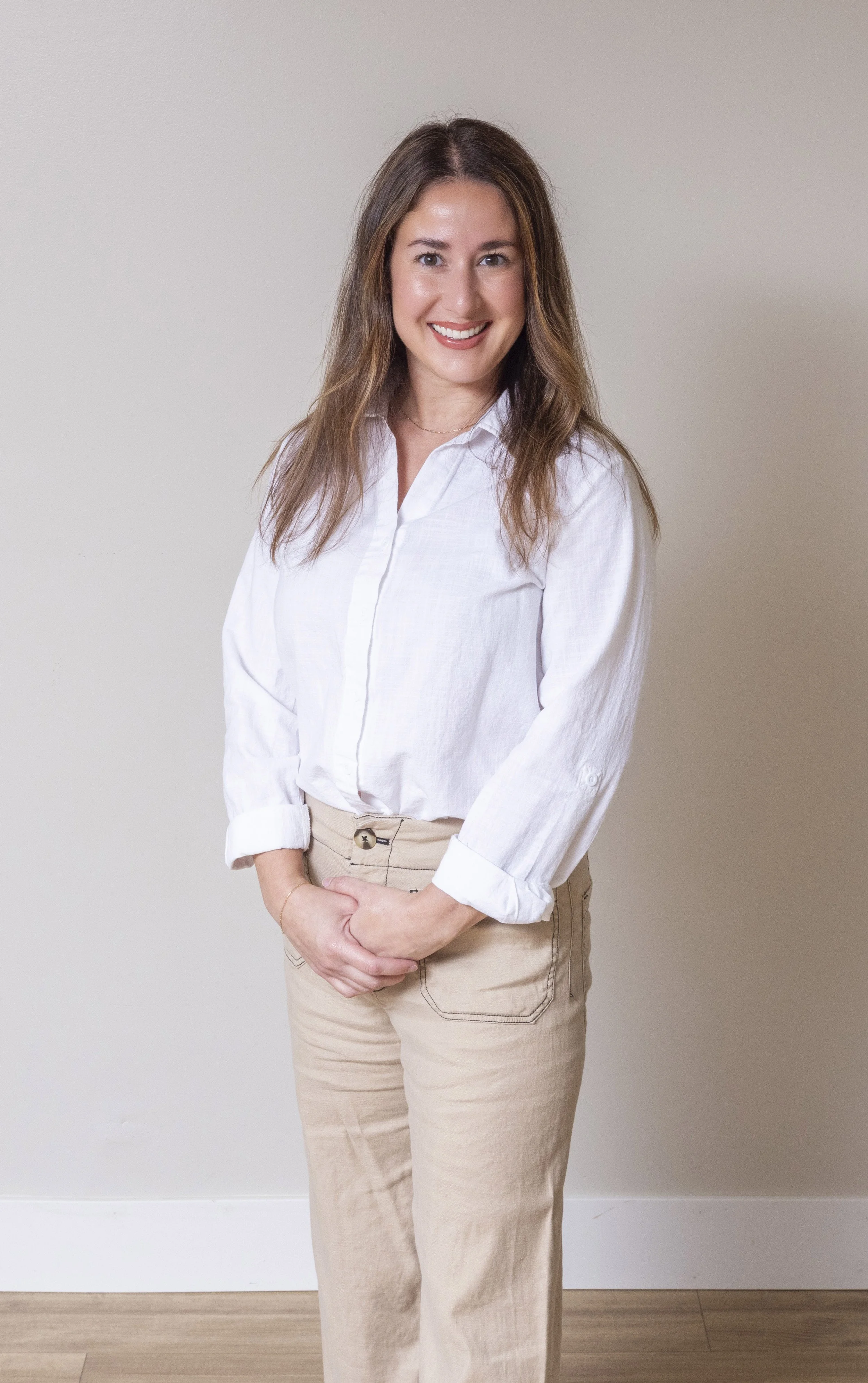 A Caucasian woman with long brown hair stands smiling with wearing a white button down top and khaki pants