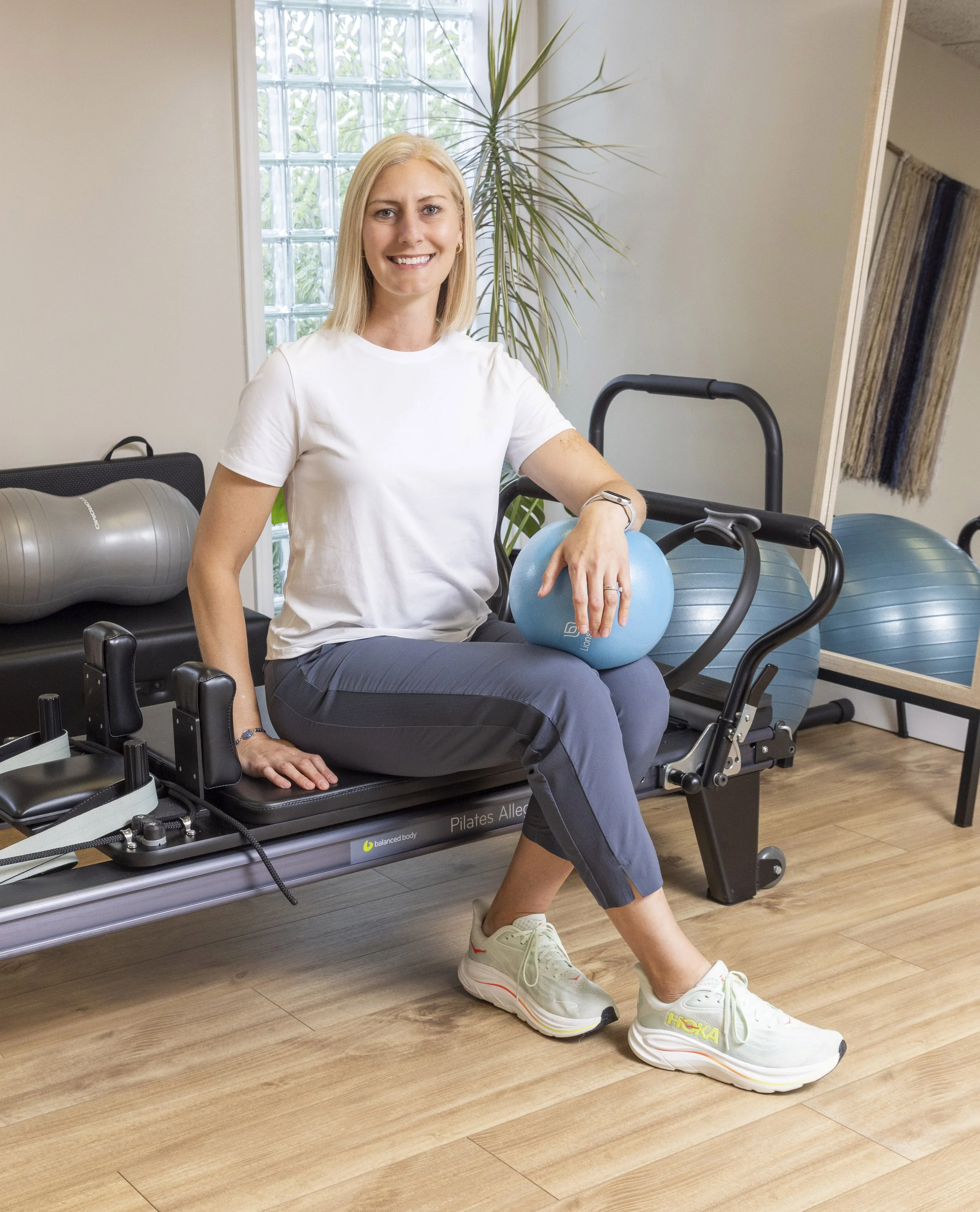 Woman sitting on a Pilates reformer at a fitness studio, holding a light blue exercise ball, wearing a white t-shirt, blue-grey pants, and white sneakers, with exercise equipment and a mirror visible in the background.