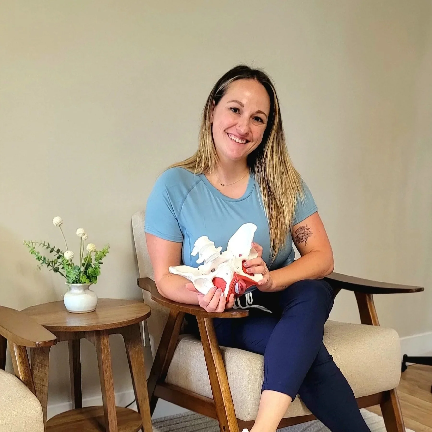 A smiling blonde woman wearing a light blue top and navy blue bottoms sits in the lobby of the Queen City Pelvic Health office holding a pelvis model.