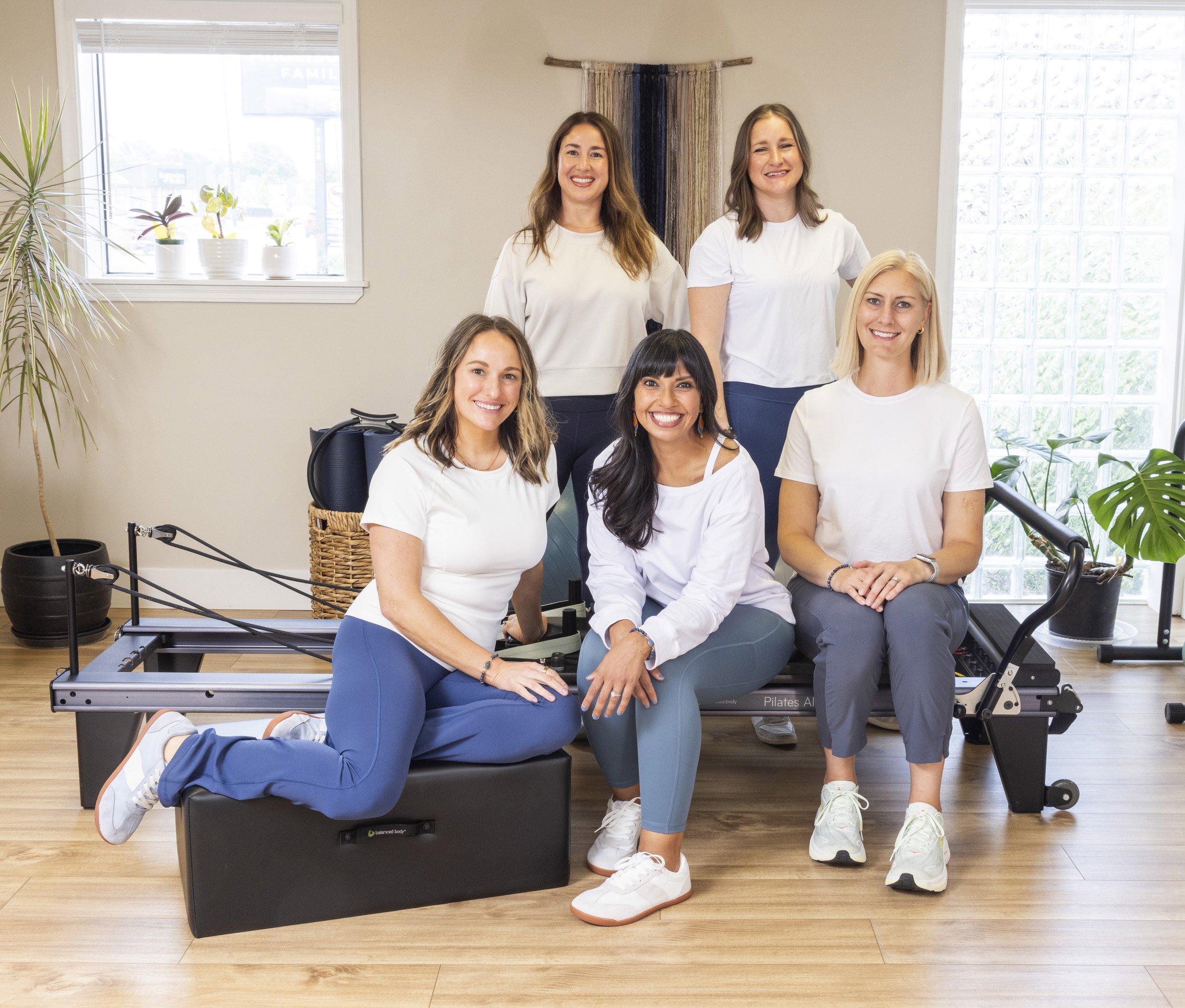 Six women wearing athletic apparel sit and stand smiling around a Balanced Body Allegro I Reformer.