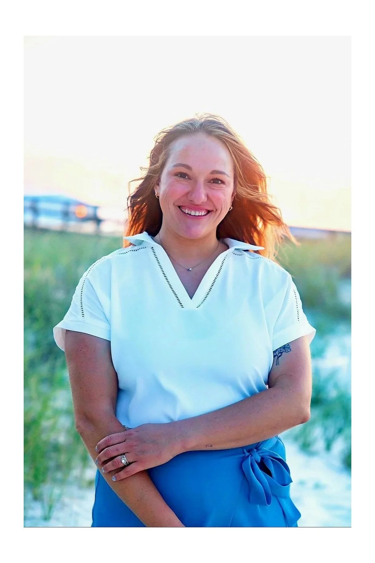 A woman wearing a white top and blue bottoms stands outdoors in a beach setting while smiling