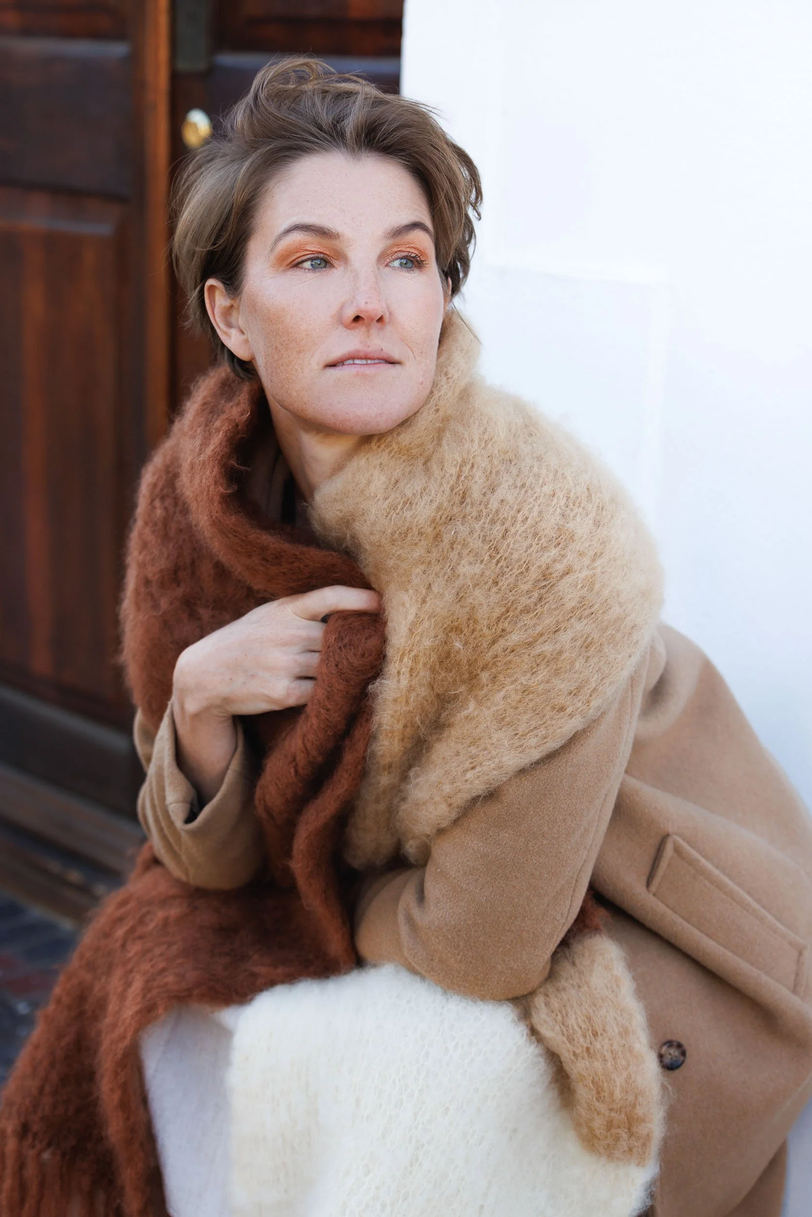 A woman with short brown hair and blue eyes wearing a beige coat and a brown and beige scarf, looking into the distance. She is sitting outdoors with a wooden structure in the background.