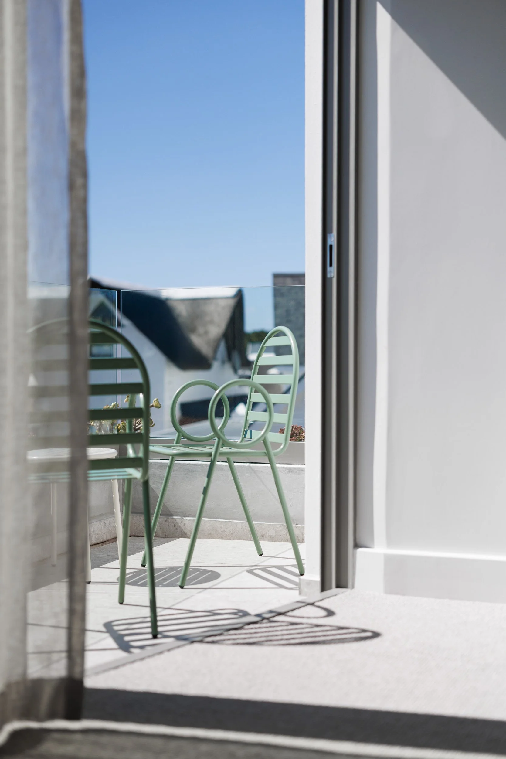 View through sheer curtain to balcony with two sage green sculptural metal chairs in bright sunlight