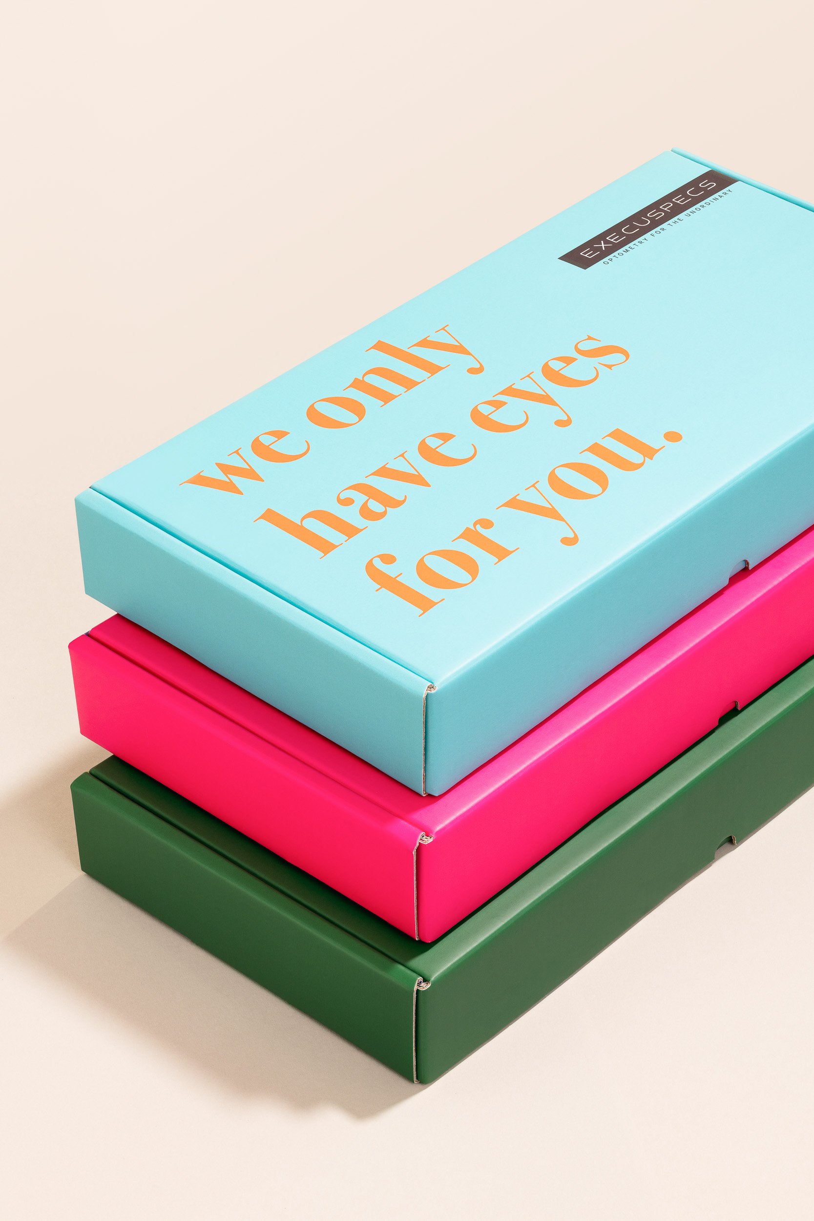 Stack of colorful boxes with motivational messages, the top box is light blue with orange text saying "we only have eyes for you" and has a black label that reads "EXECUSECTS OPTOMETRY FOR THE JOURNEY". Below are pink, green, and another pink box.