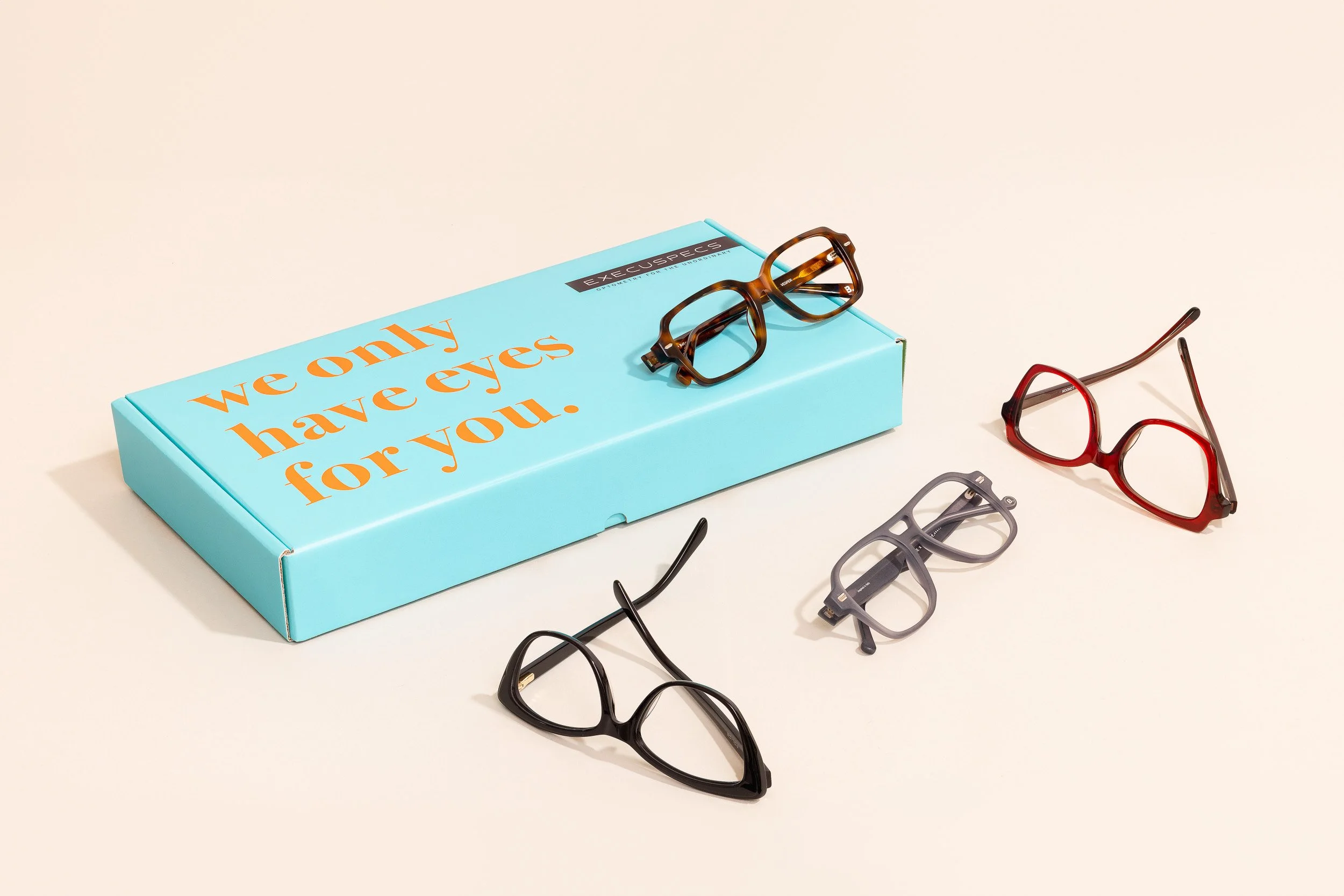 Several pairs of glasses, including black, red, and gray frames, are scattered on a white surface next to a light blue box with the phrase "we only have eyes for you" printed in orange and black.
