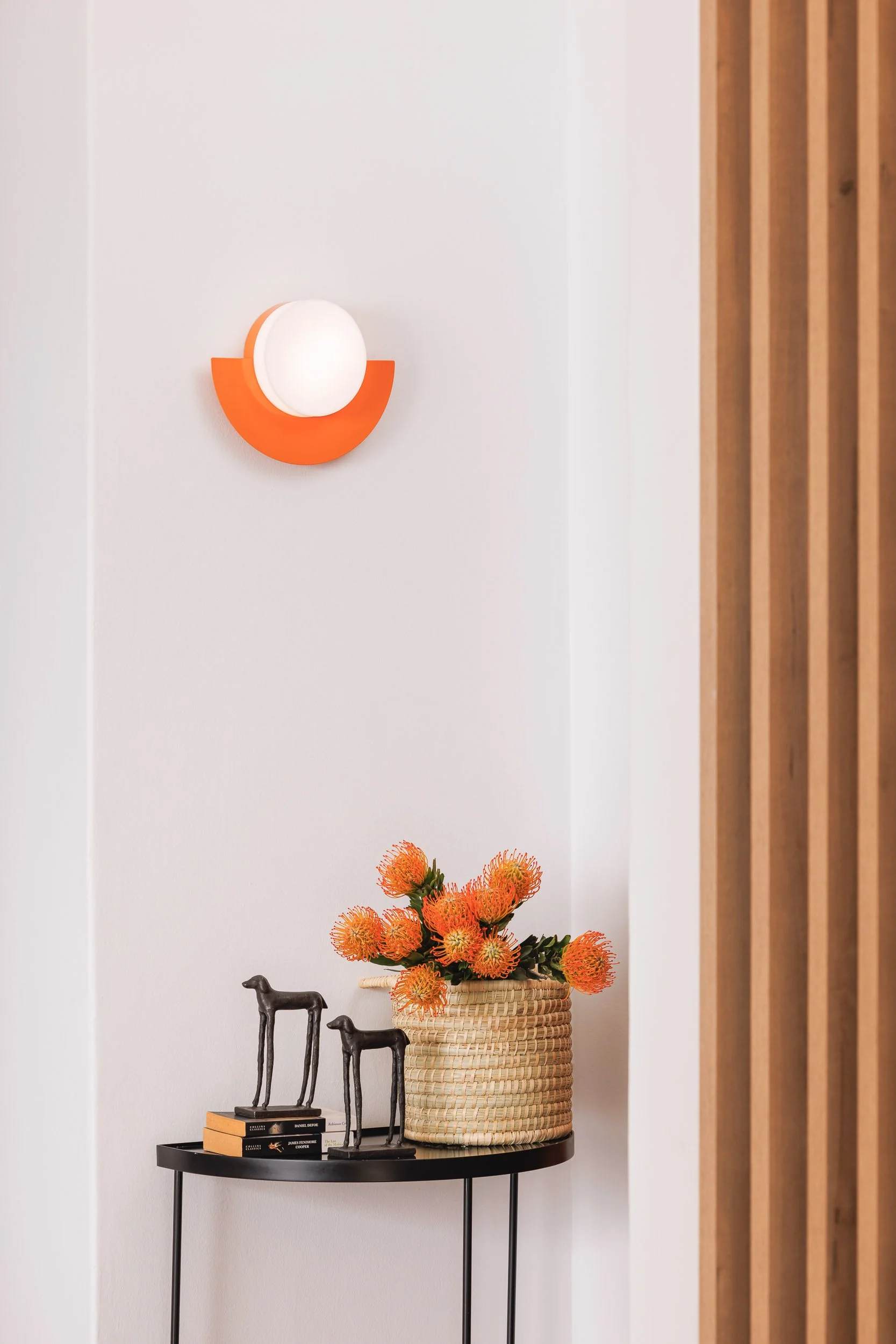 A black round console table with three books and three metal dog sculptures on top, a large woven basket with bright orange flowers, a modern white and orange wall sconce, and a wooden slat wall on the right side.