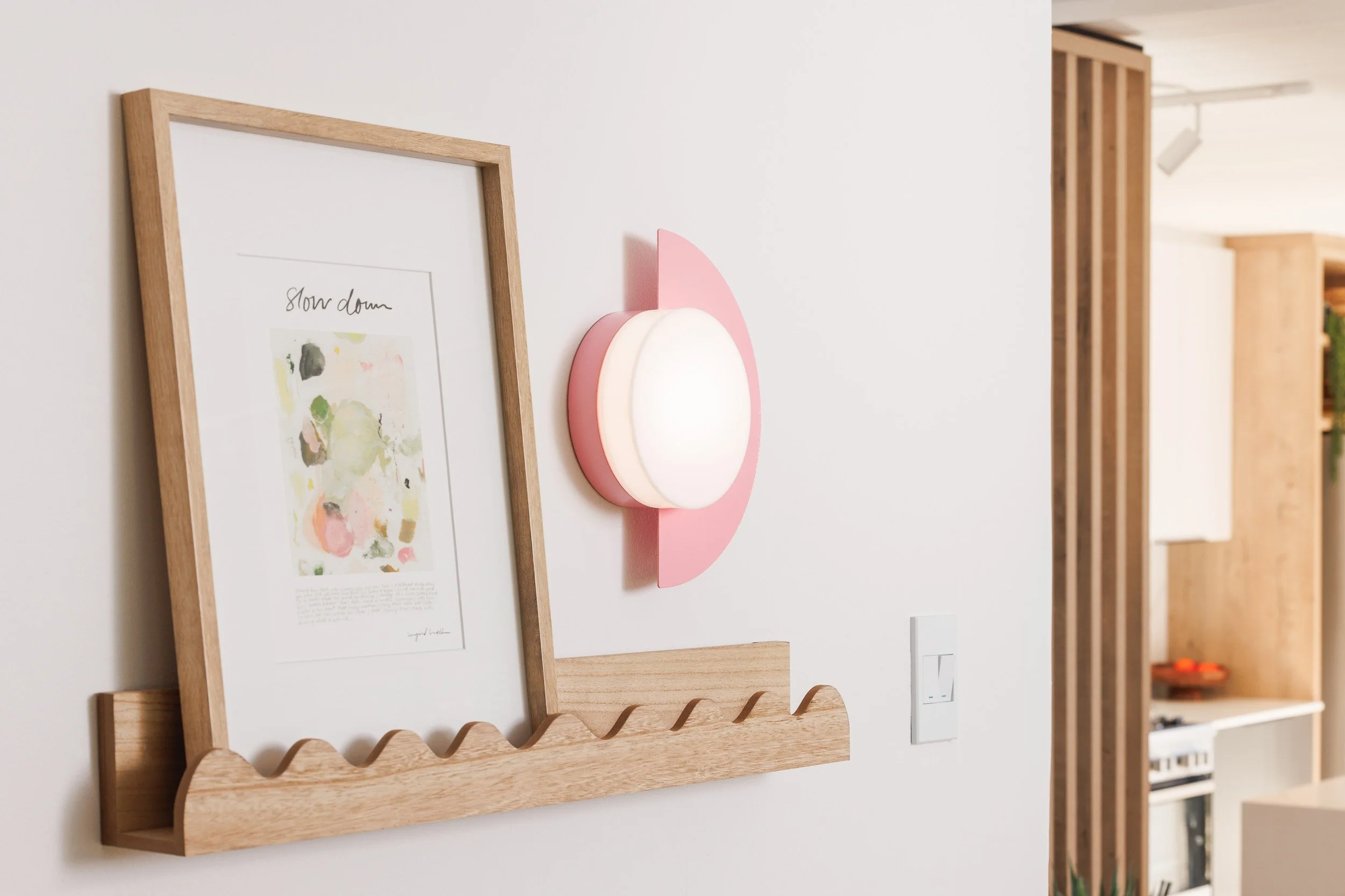 Interior view of a modern living space with a white wall, a wooden picture frame with an abstract watercolor art, and a pink and white wall lamp with geometric shapes.