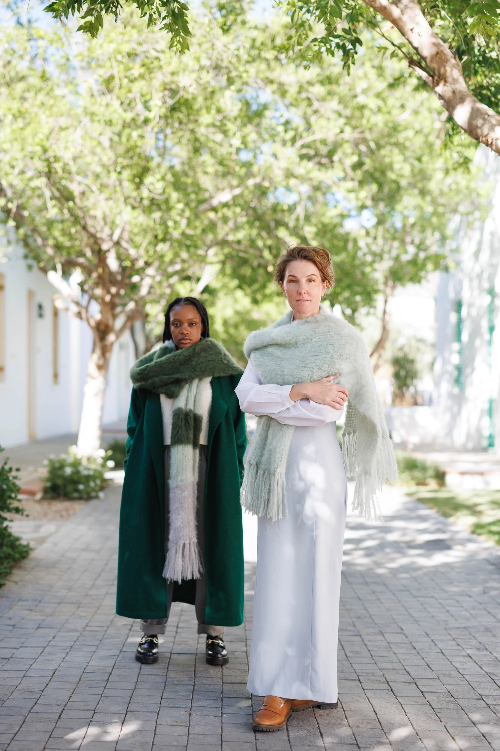 Two women standing outside on a paved walkway with green trees in the background. One woman has short hair and is wearing a white dress with a light green shawl, and the other has braided hair and is dressed in dark green with a multicolored scarf.