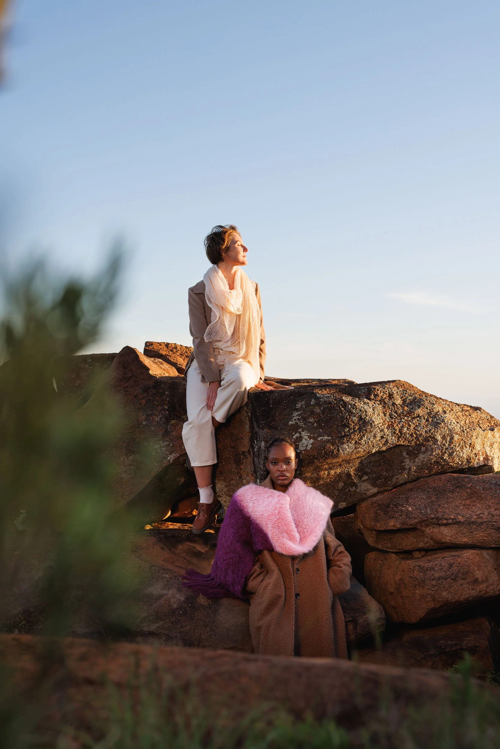  women posing on large rocks outdoors during sunset. The woman at the top has short hair, is dressed in a light-colored jacket and scarf. The woman on the bottom has dark skin, short hair, wearing a pink scarf, seated on the rocks. 