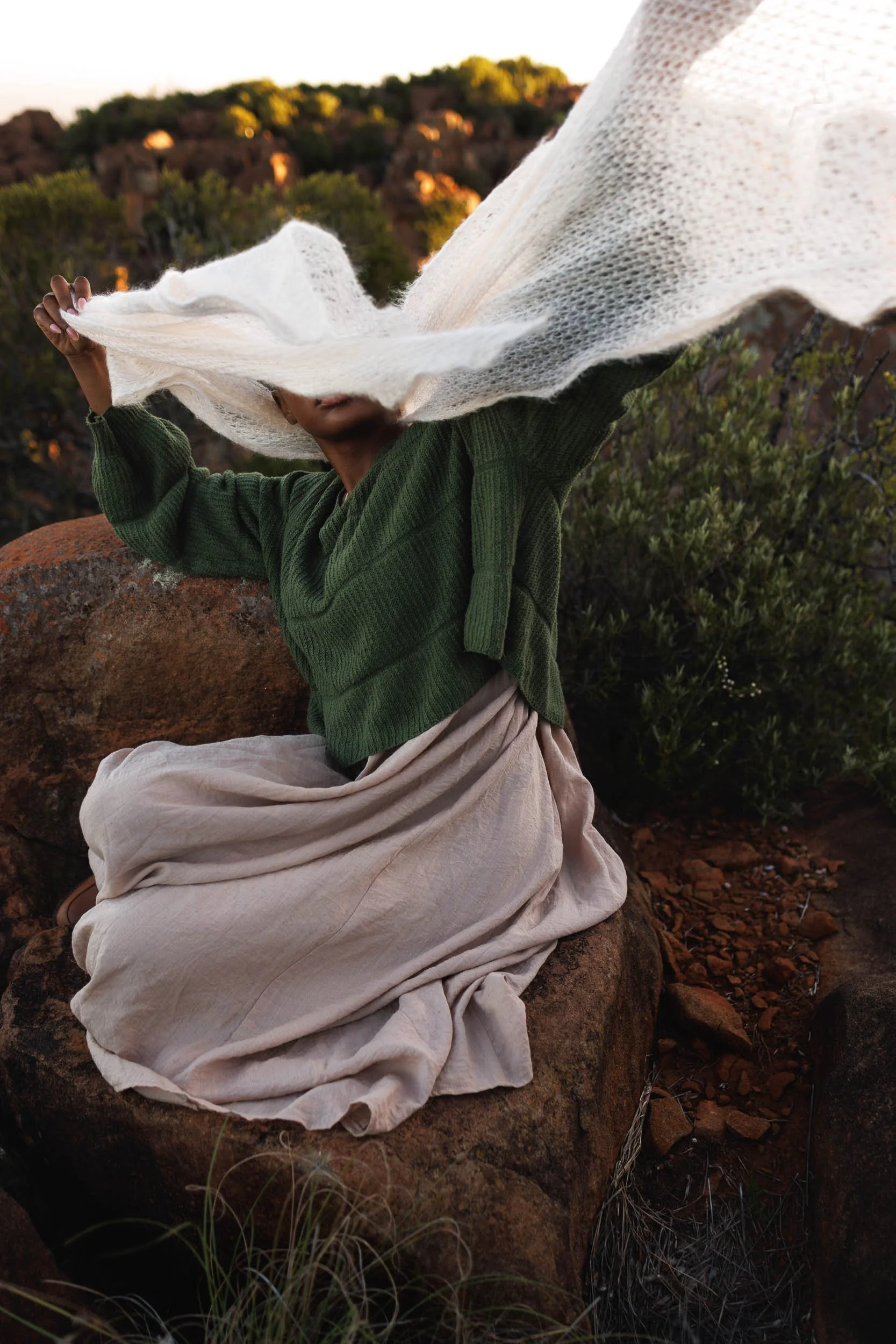Person sitting on a large rock, holding a white cloth or scarf, with a background of hills and shrubbery during sunset.